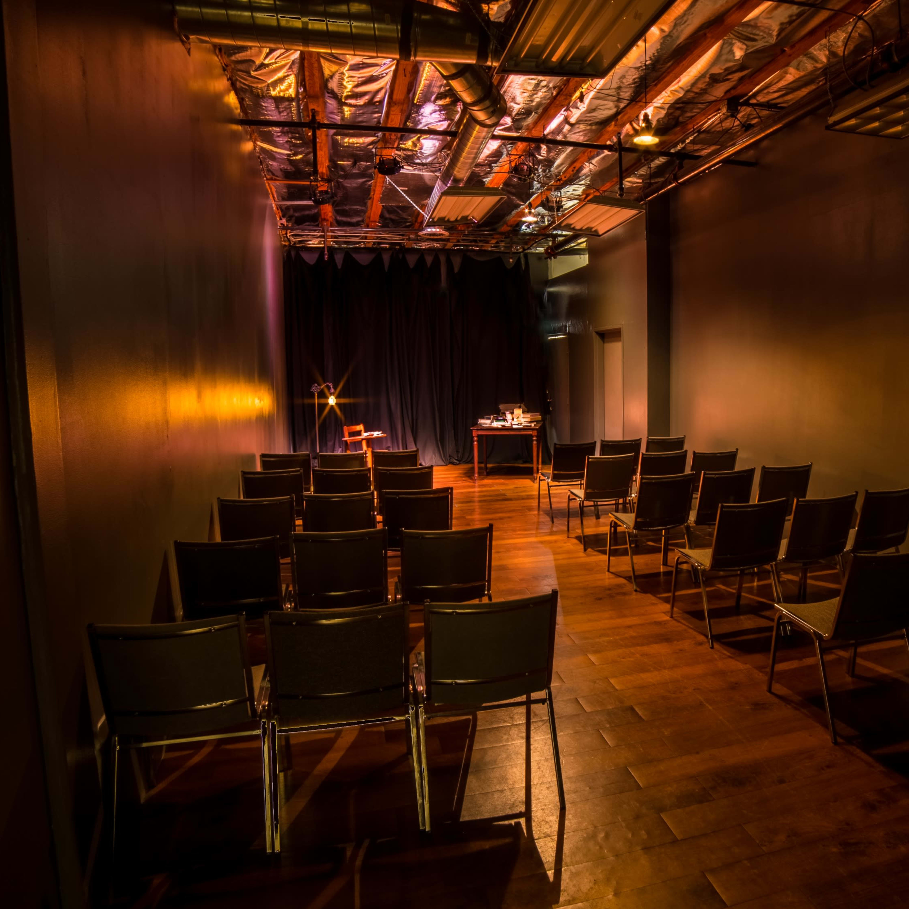 A dimly lit auditorium with rows of metal chairs facing a small stage and a table at the front.