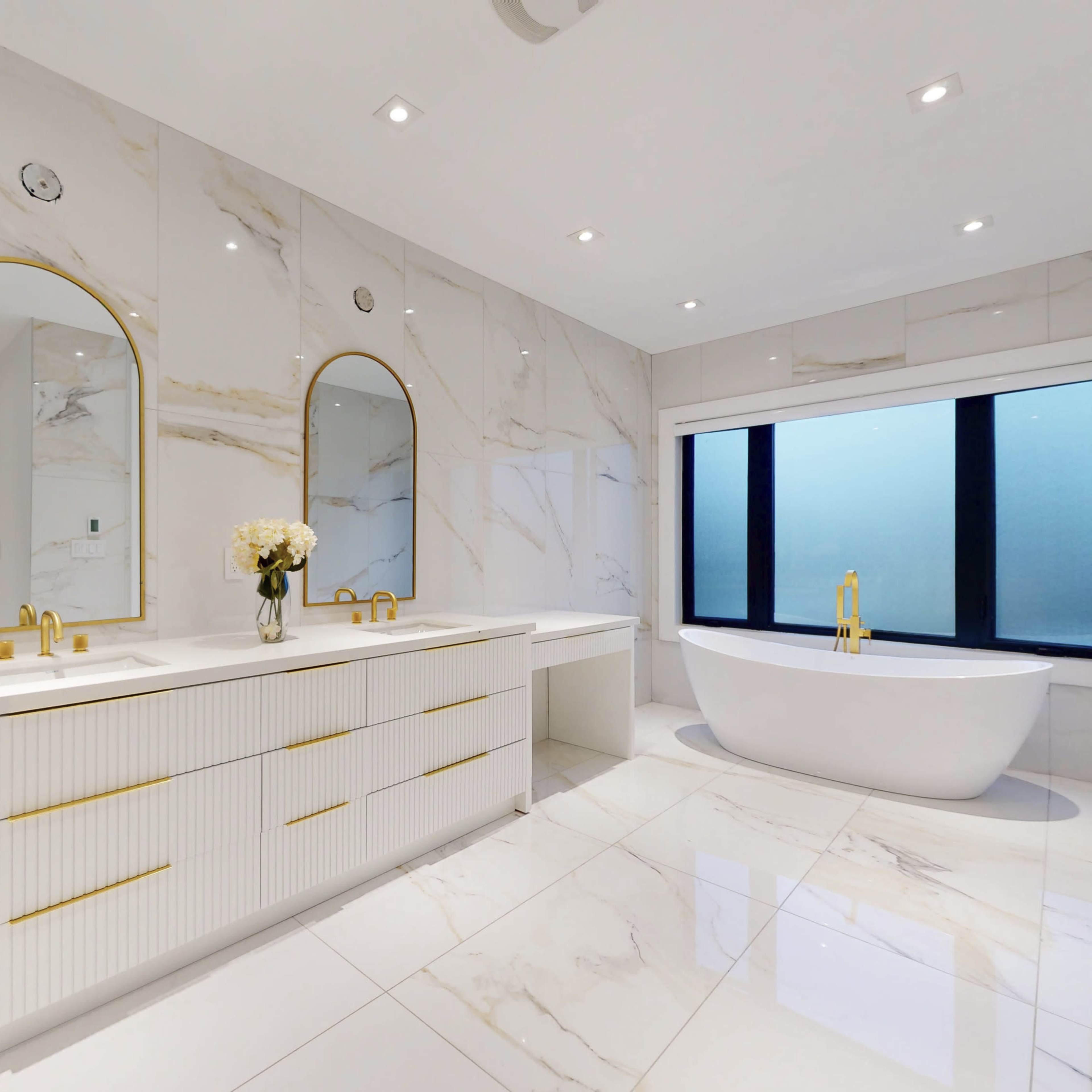 The bathroom features dual sinks with round mirrors, a freestanding tub, and large windows, all set against a backdrop of marble-tiled walls and floors.