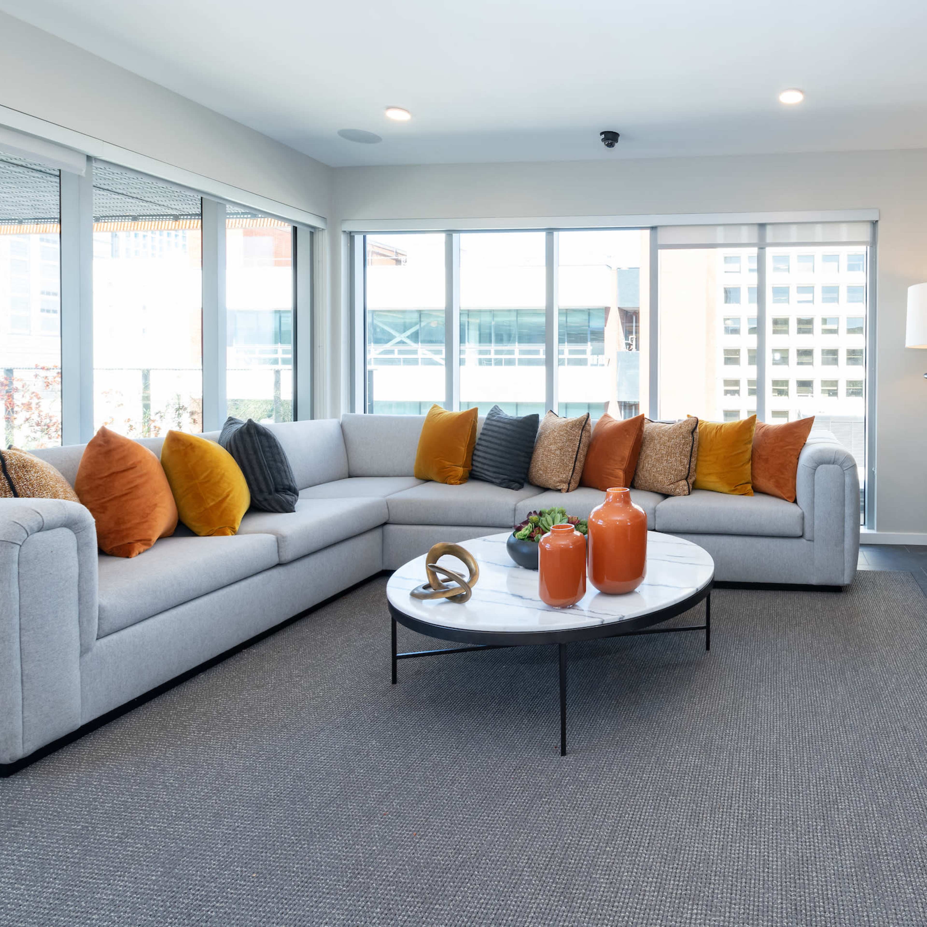 A modern living room with a large gray sectional sofa adorned with colorful pillows, a circular coffee table, and large windows providing natural light.