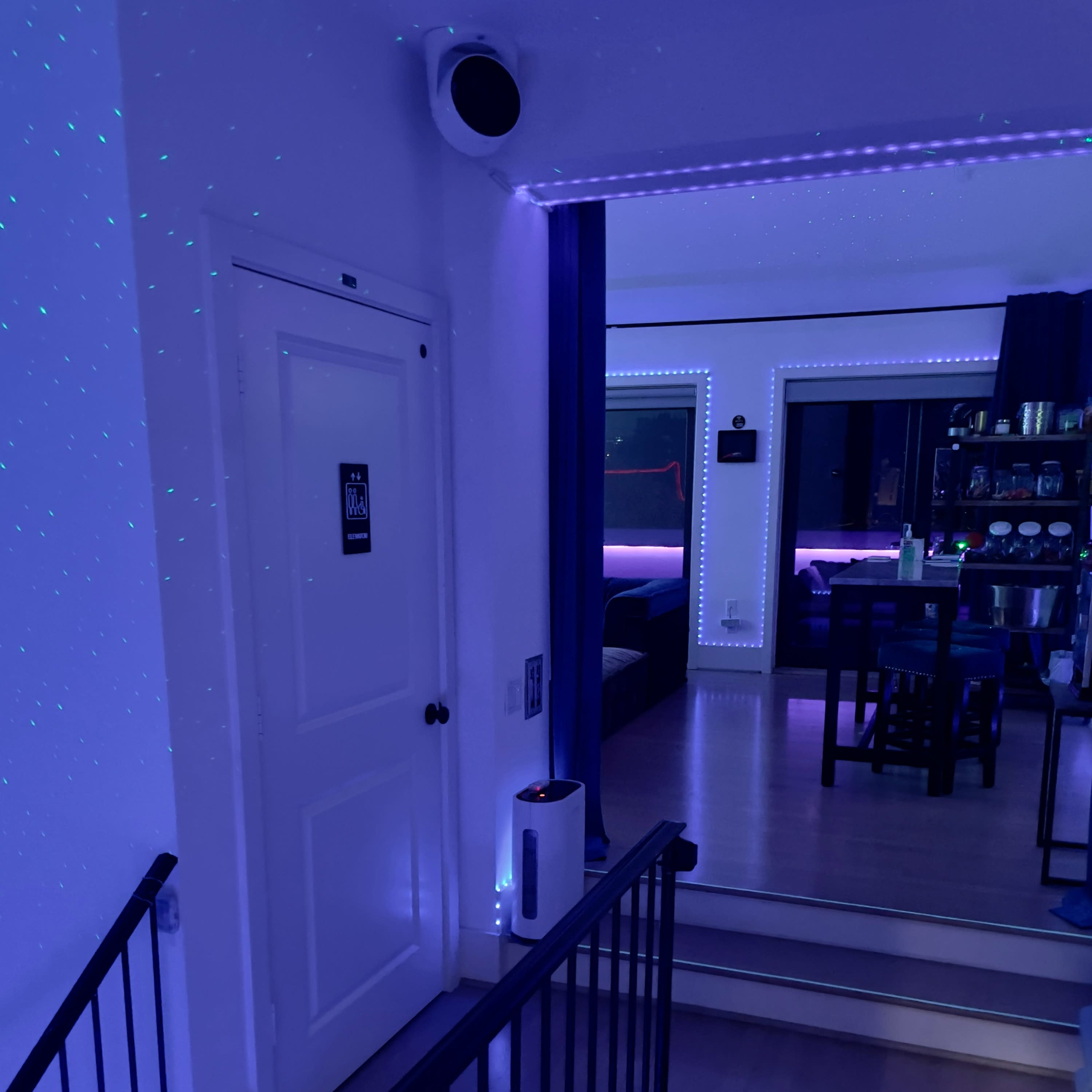 A staircase leading to a hallway that connects to a brightly lit room with blue lighting and various furnishings.