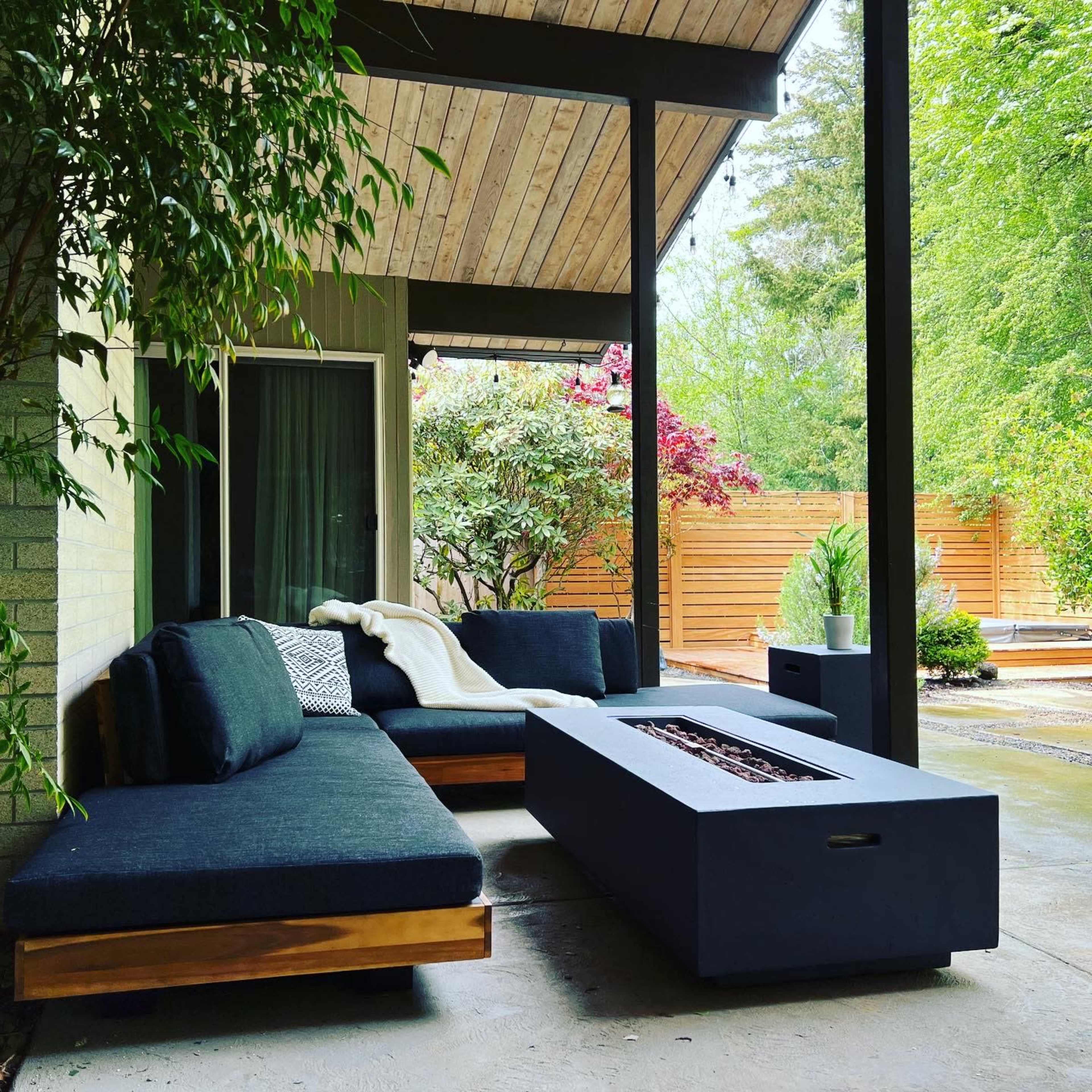 A modern outdoor seating area featuring a sectional sofa, a square fire pit, and lush greenery in the background.