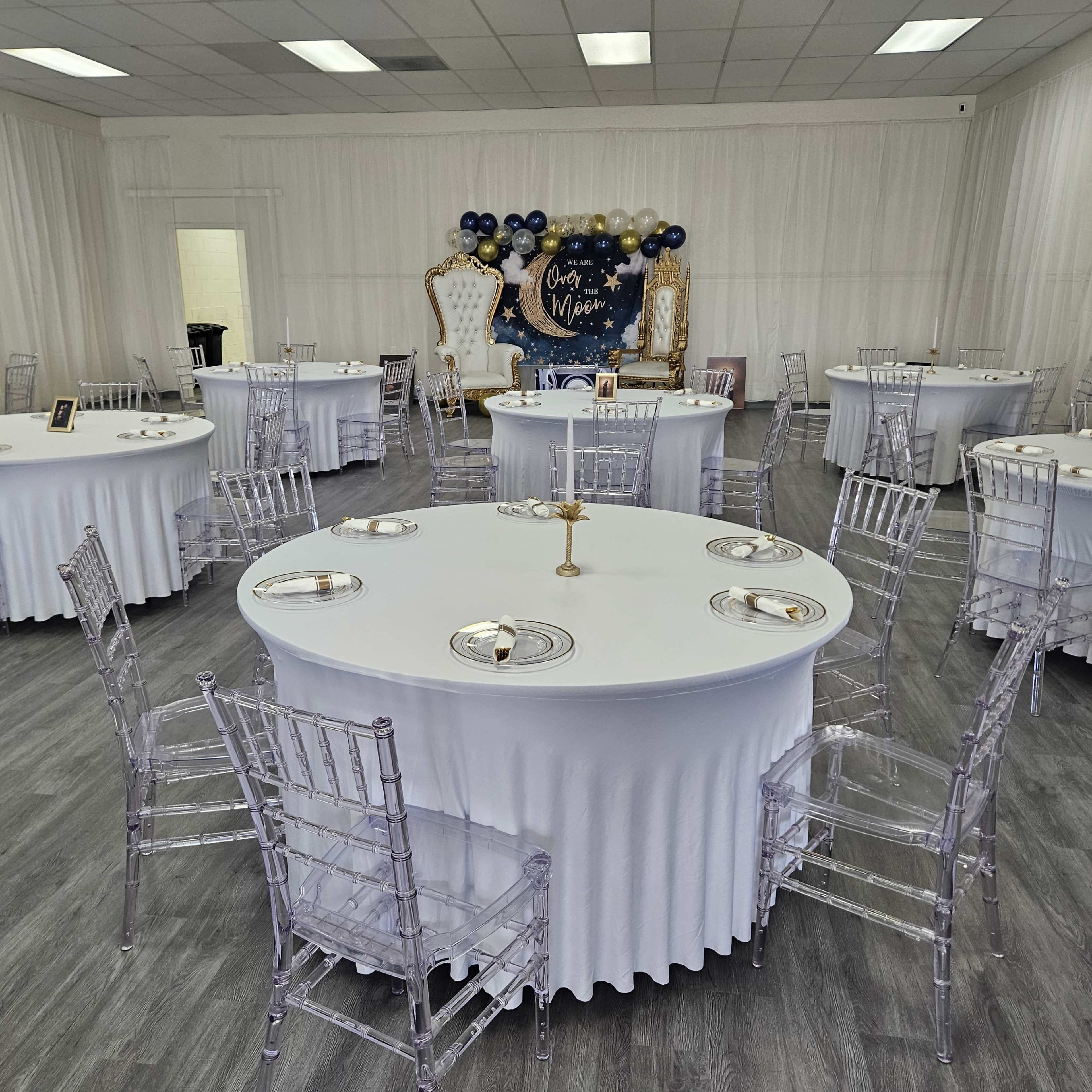 A spacious event room arranged with several round tables covered in white tablecloths and surrounded by clear acrylic chairs, set for a formal gathering.