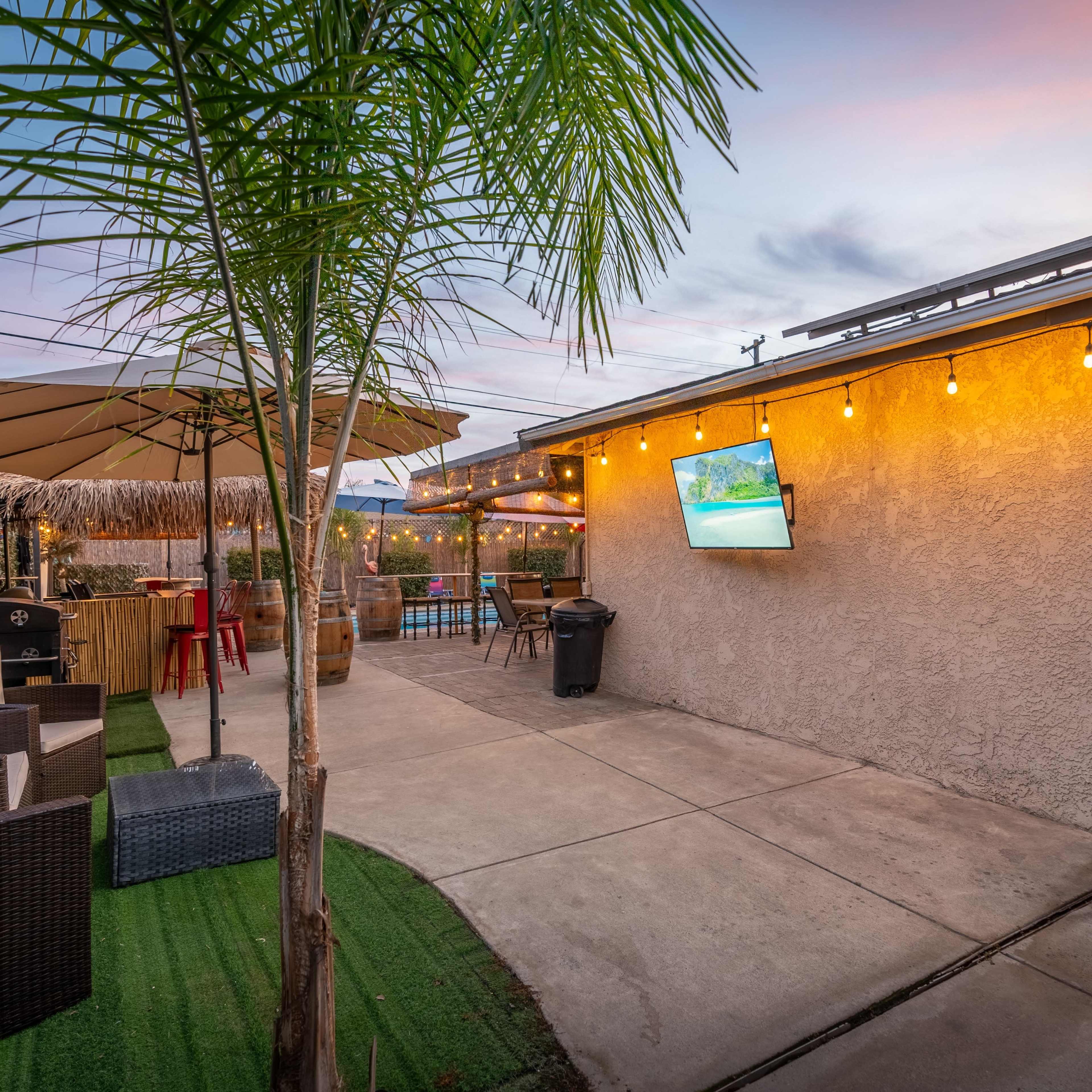A backyard patio area featuring a seating arrangement, a large umbrella, string lights, and a mounted TV on the wall, all set against a backdrop of a swimming pool.