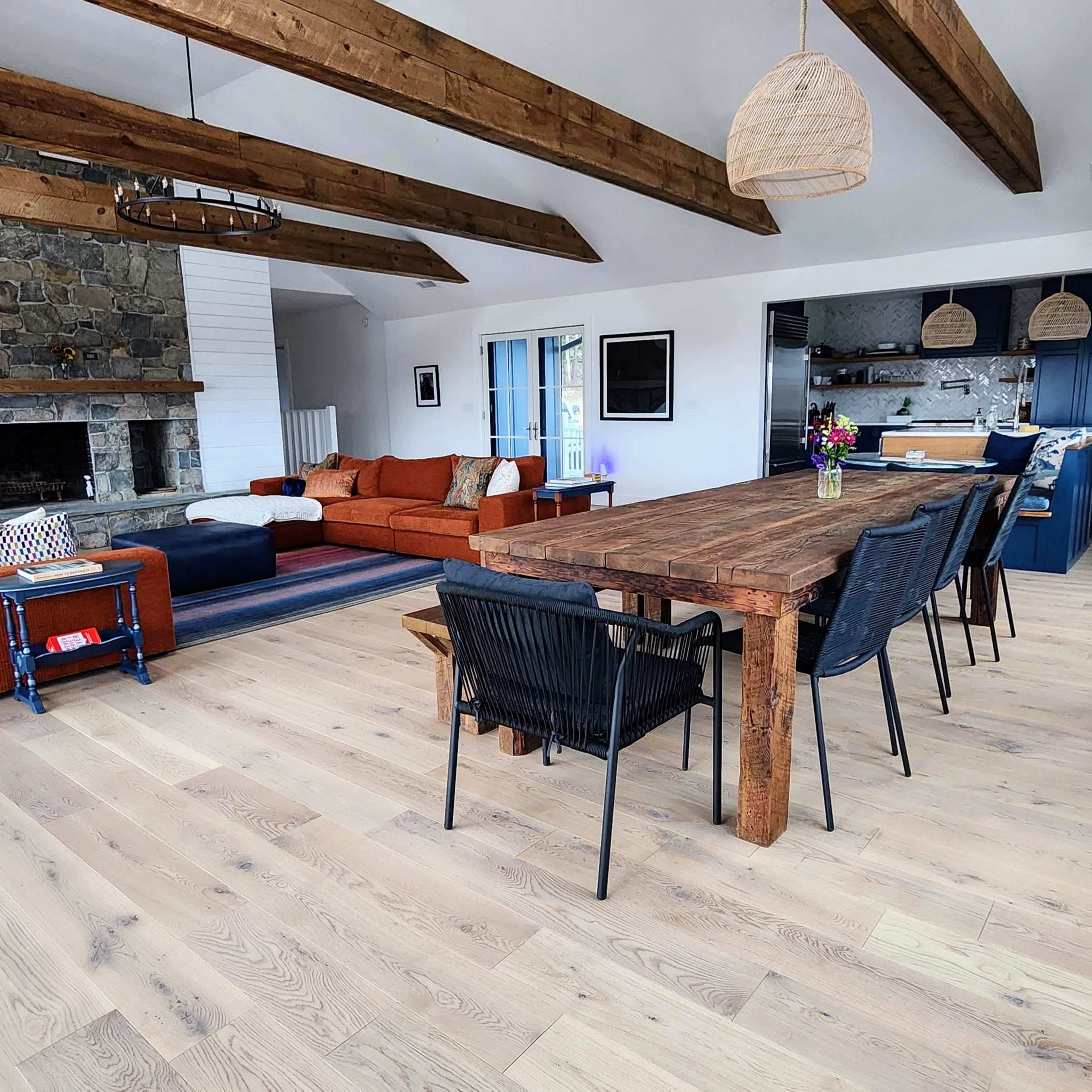 A spacious living and dining area with wooden beams, a stone fireplace, an orange sectional sofa, a long wooden dining table, and a well-lit kitchen in the background.