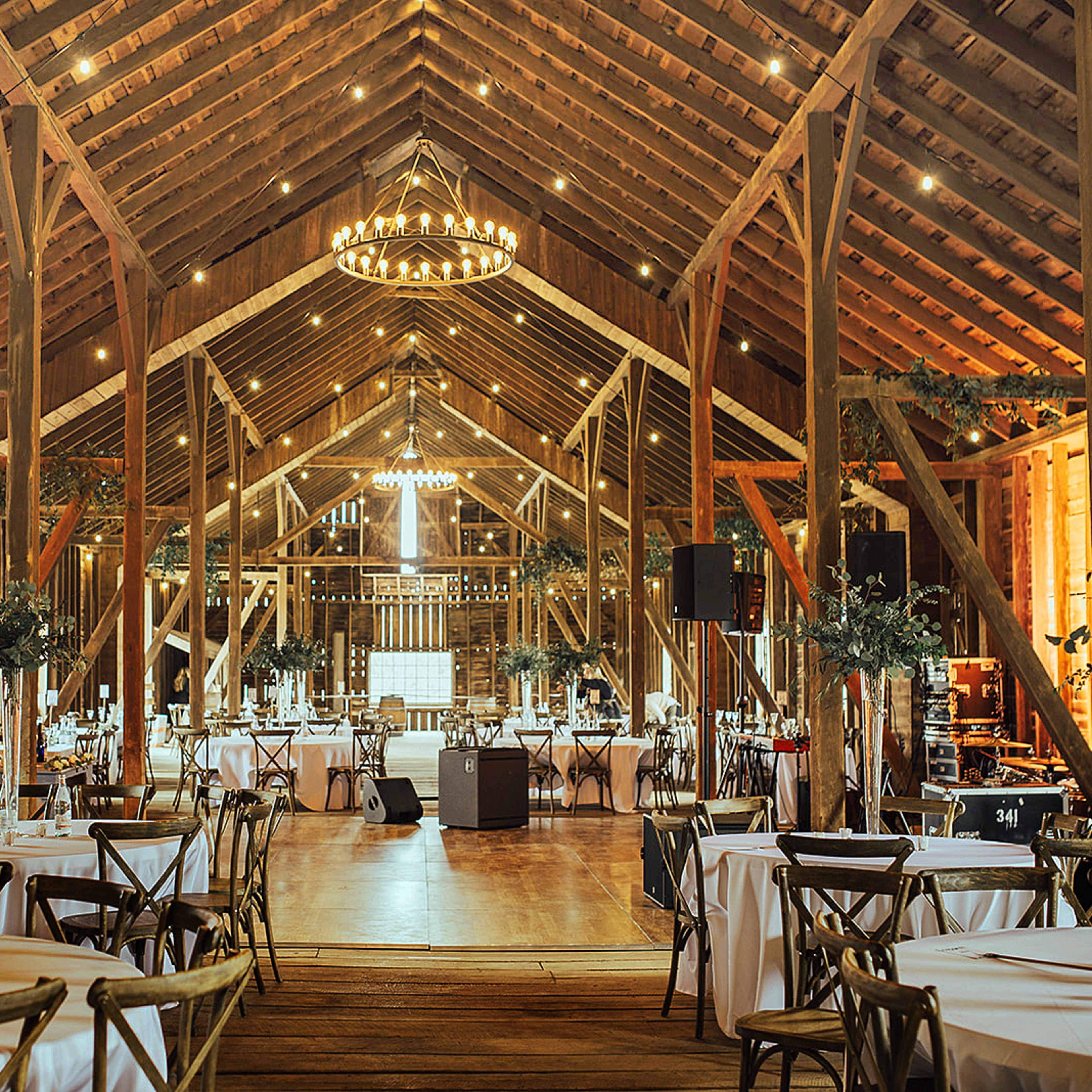 The interior of a spacious barn features round tables with white tablecloths, rustic wooden beams, and chandeliers hanging from the high ceiling.