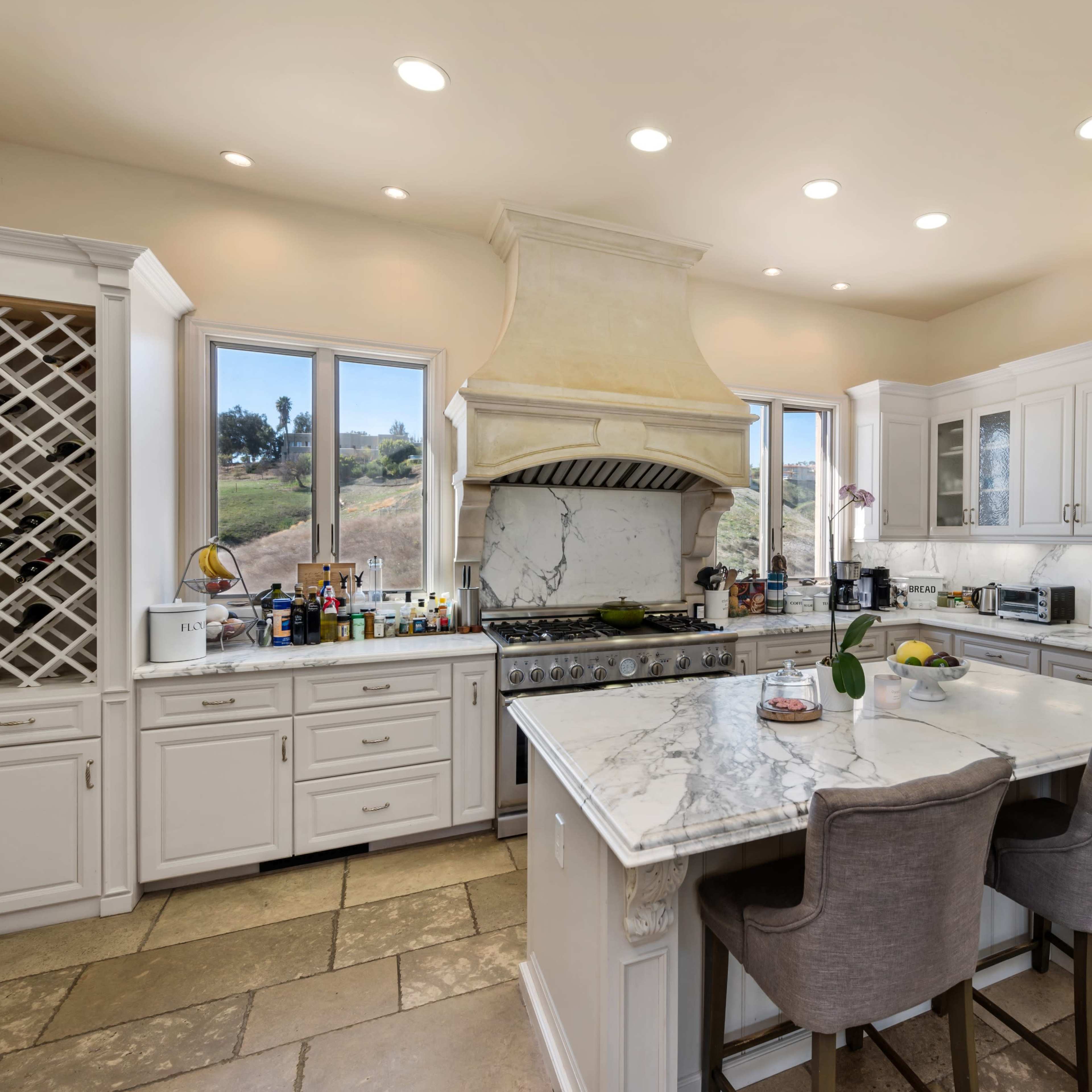 A spacious kitchen features white cabinetry, a marble countertop island, and large windows overlooking a scenic landscape.