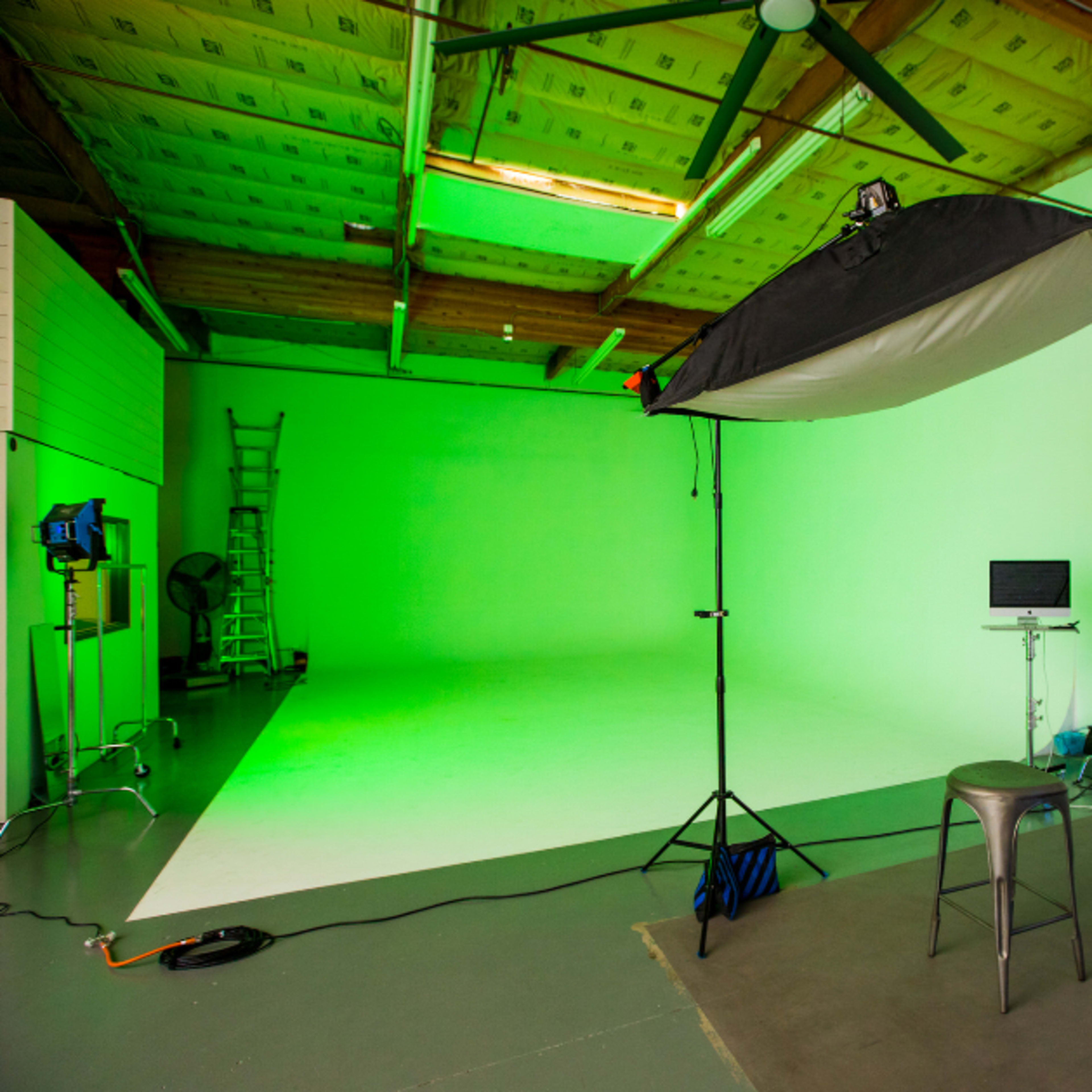 A photography studio features a green backdrop, a lighting setup, a stool, and various equipment arranged in the space.