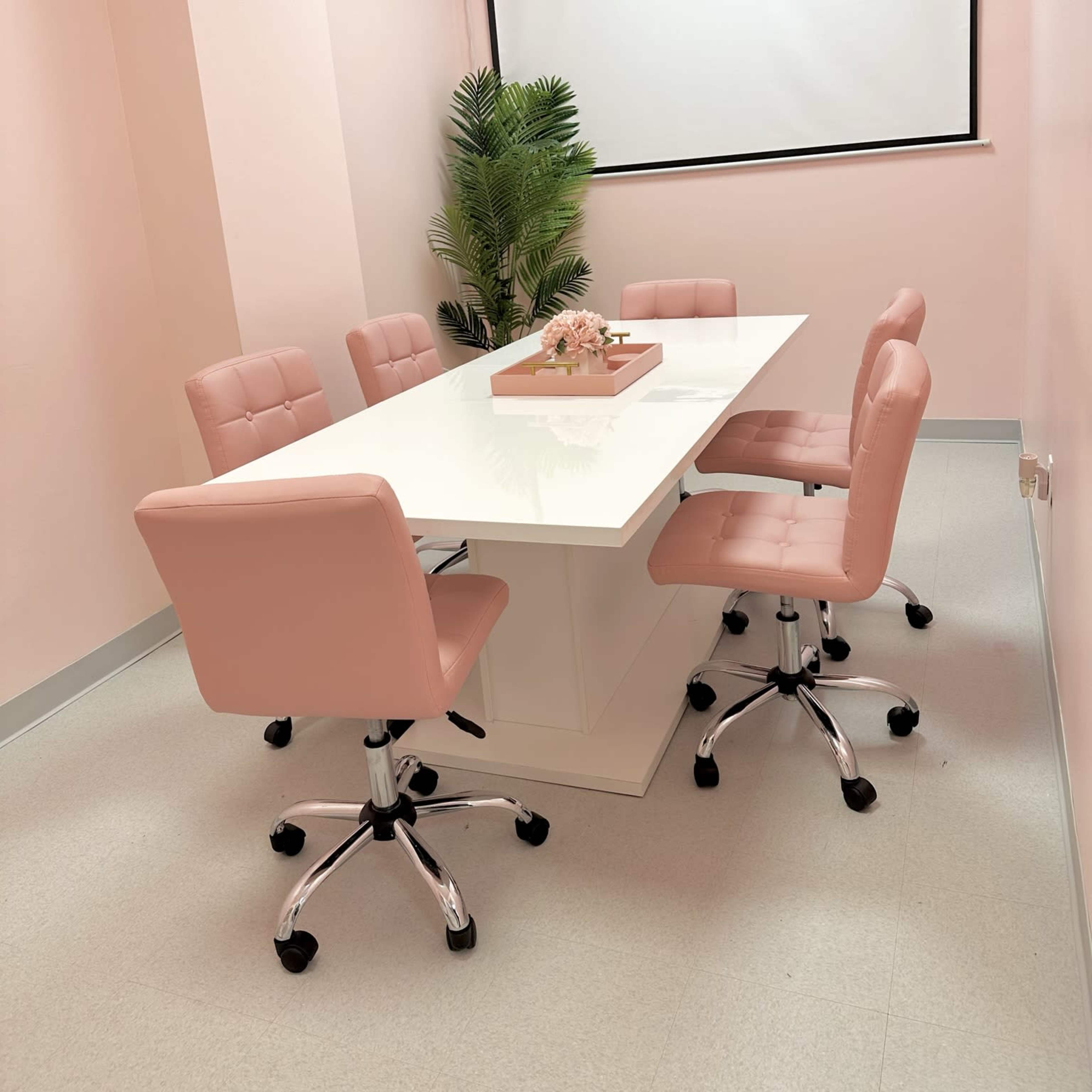 A conference room features a white table surrounded by pink chairs and a small decorative plant, with a projection screen at the front.