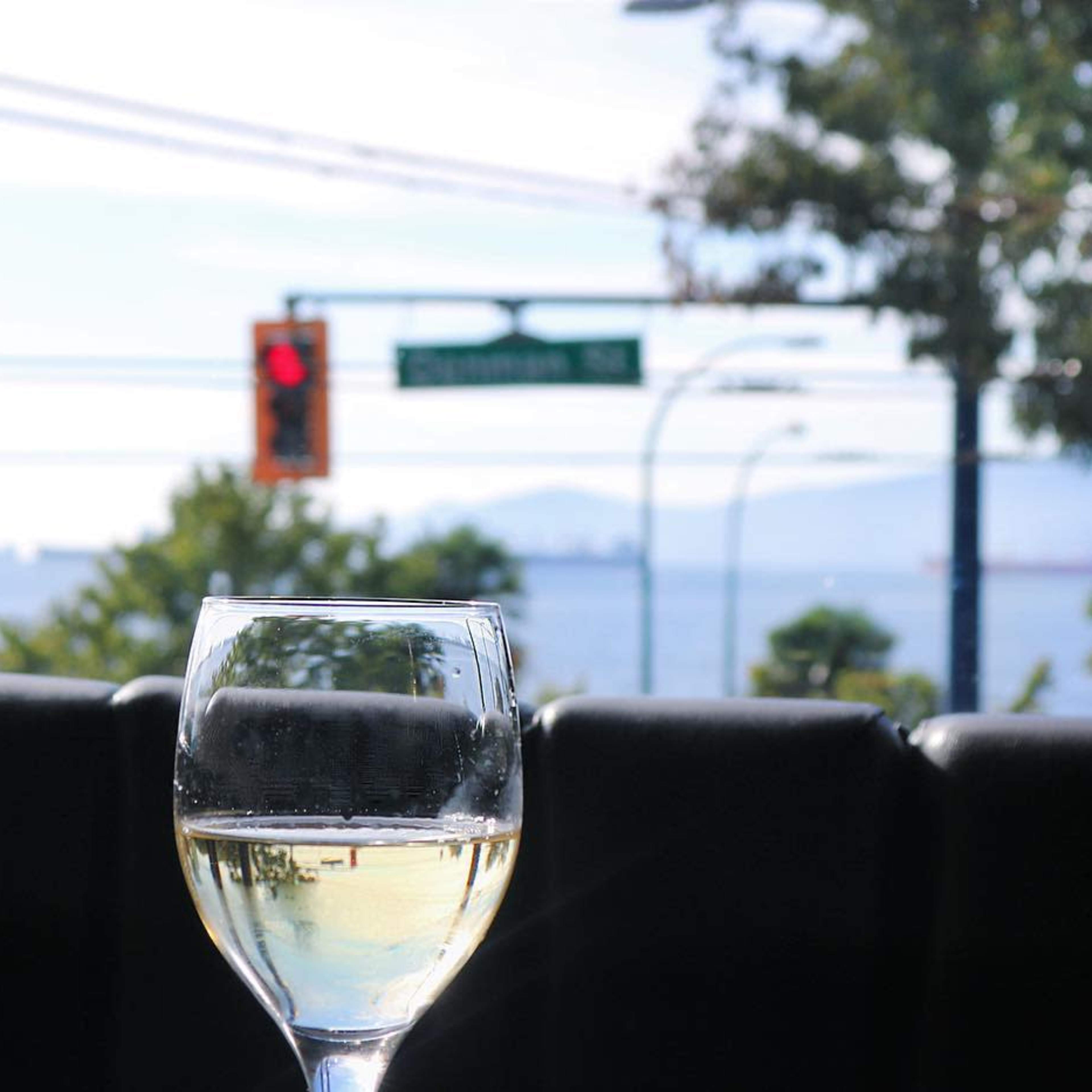 A glass of white wine sits on a table, with a view of a bay and distant ships under a clear sky.