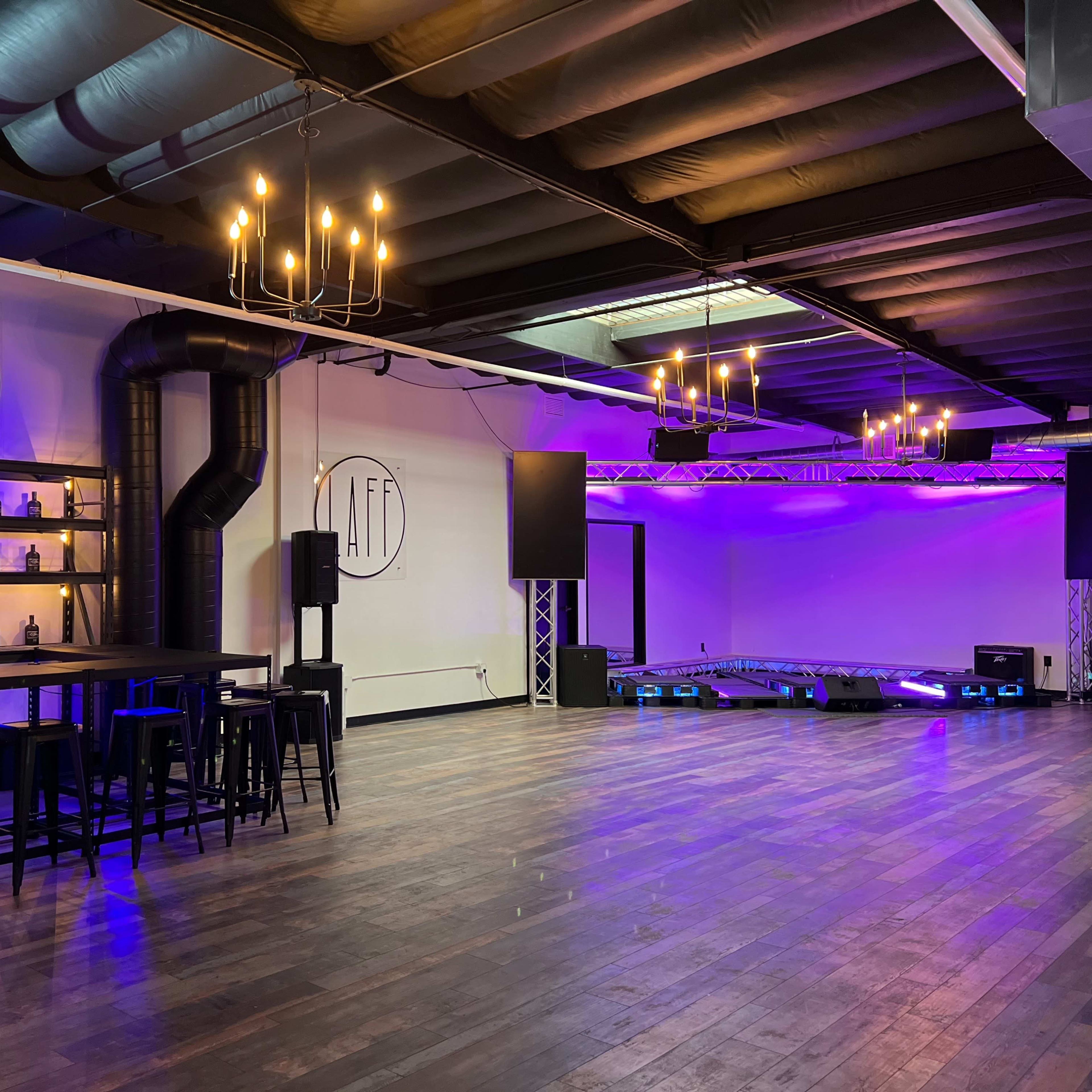 A modern event space with a wooden floor, shelving displaying bottles, and purple lighting accentuated by chandeliers.