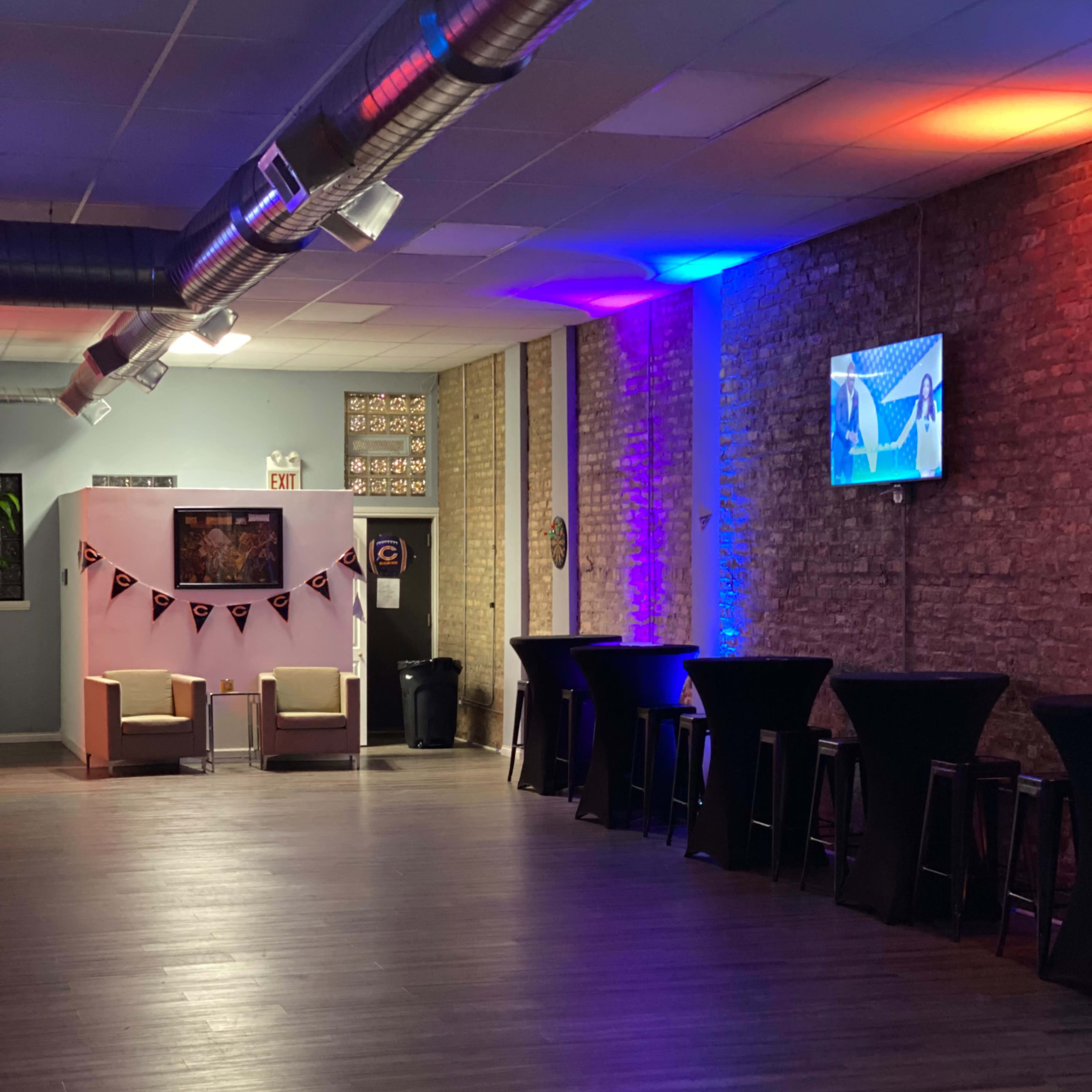 A spacious, dimly lit room featuring several tall tables, two seated areas, and a wall-mounted television displaying a scene.