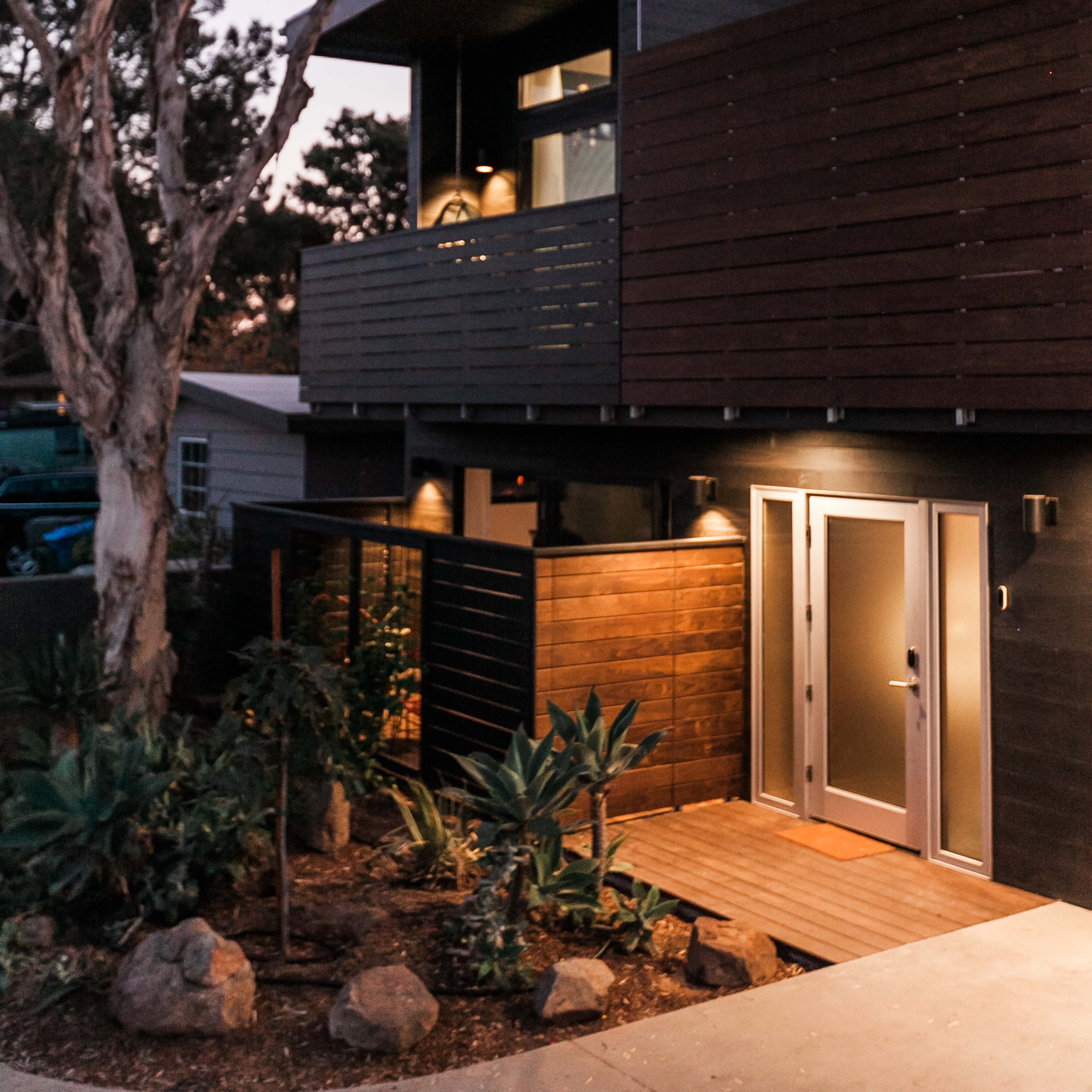 A modern two-story house features wooden exterior accents and is set among trees, with a pathway leading to its entrance.