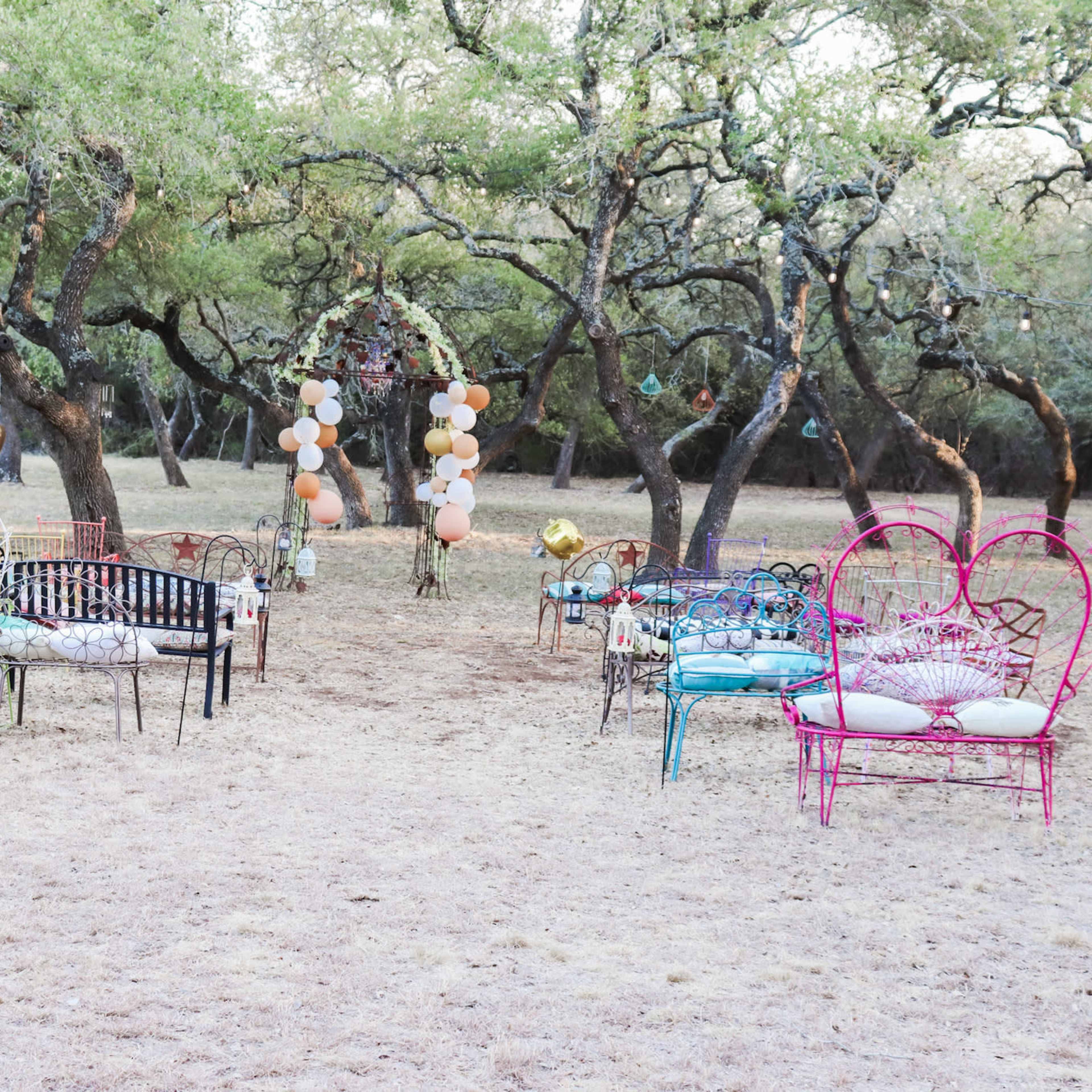 A collection of colorful metal chairs and benches is arranged in an outdoor setting beneath trees, with decorative balloons and an archway in the background.