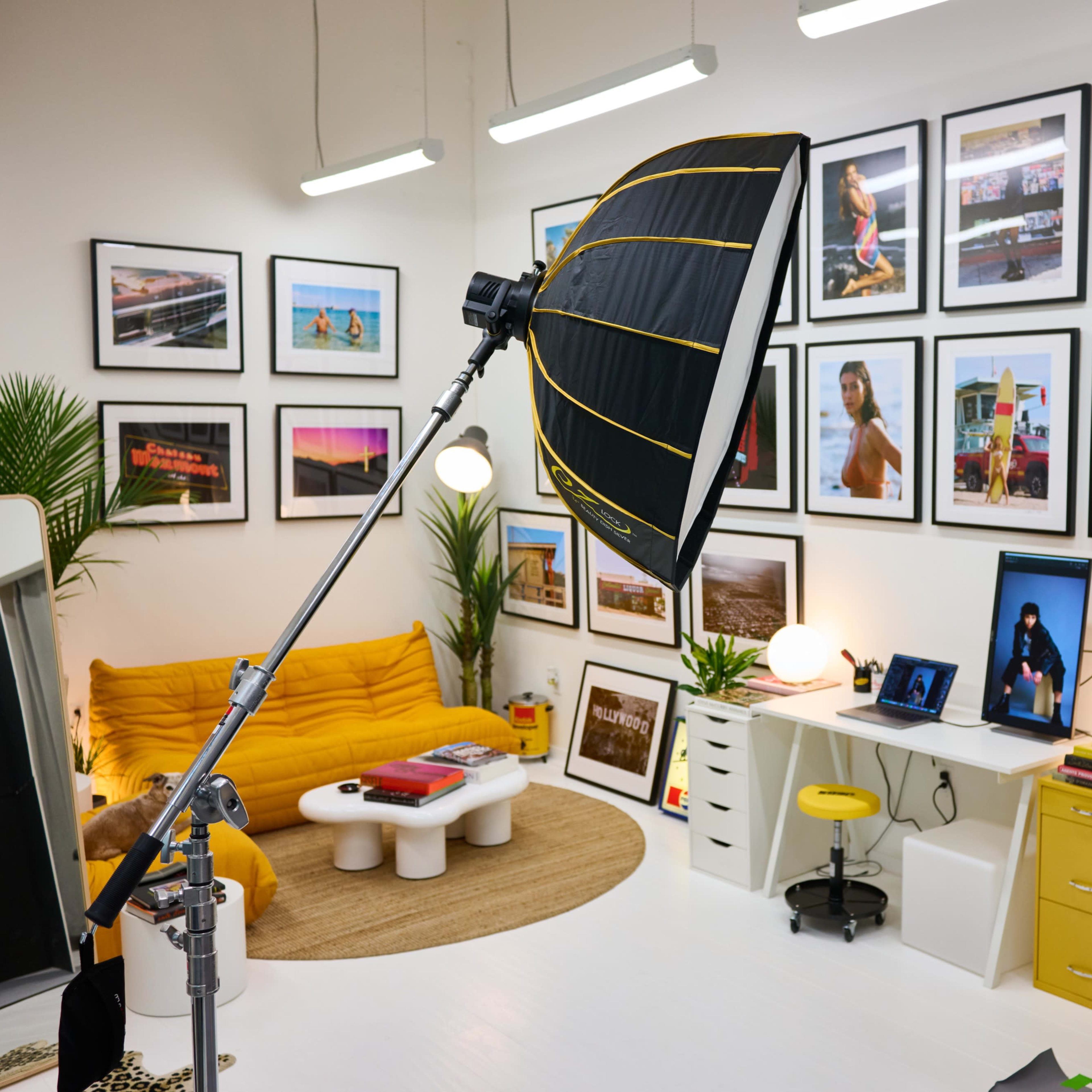A brightly lit studio space featuring a large umbrella light, a yellow sofa, a variety of framed photographs on the walls, and a workspace with a computer and yellow accents.