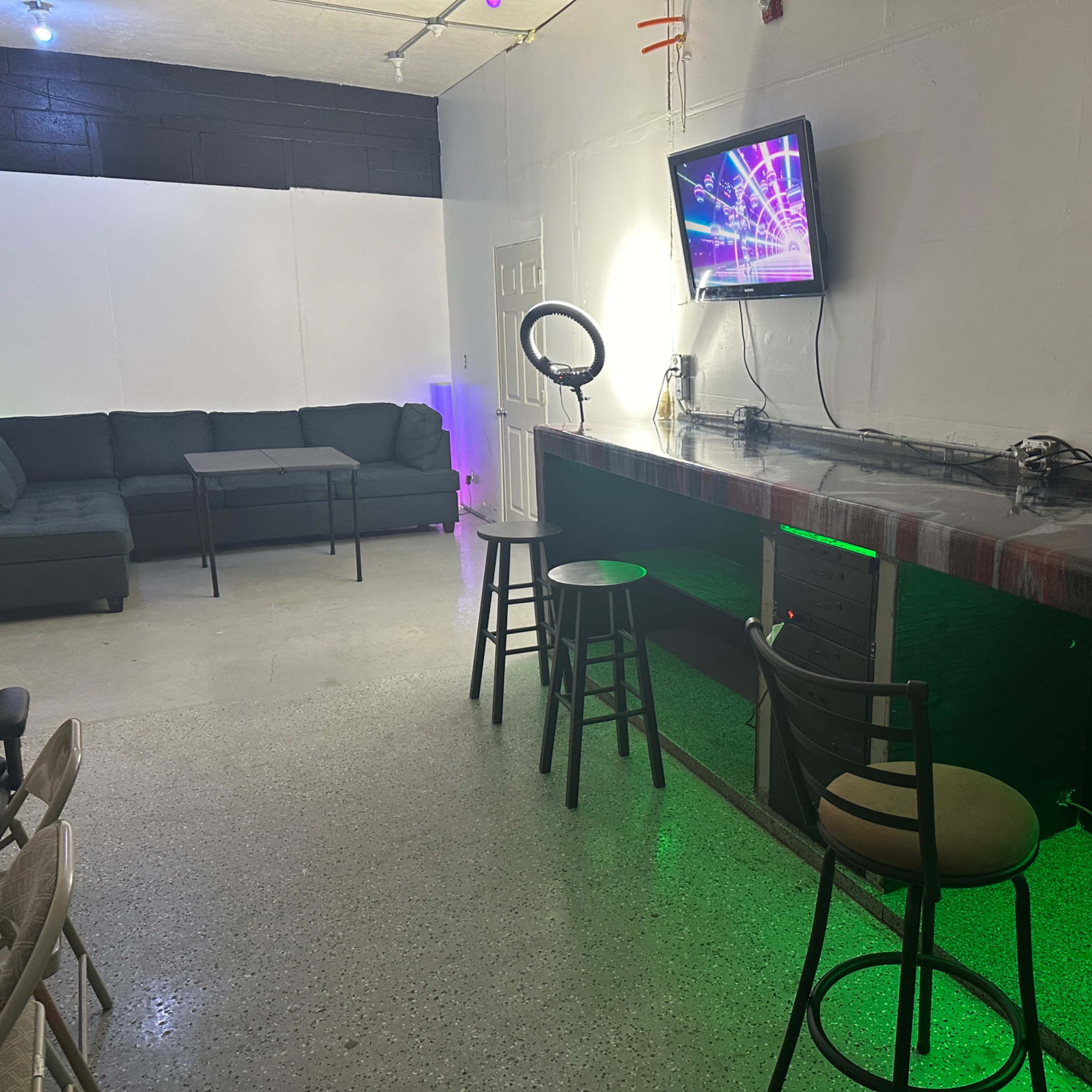A lounge area with a bar, a couch, a table, and a television mounted on the wall, illuminated by colorful lights.