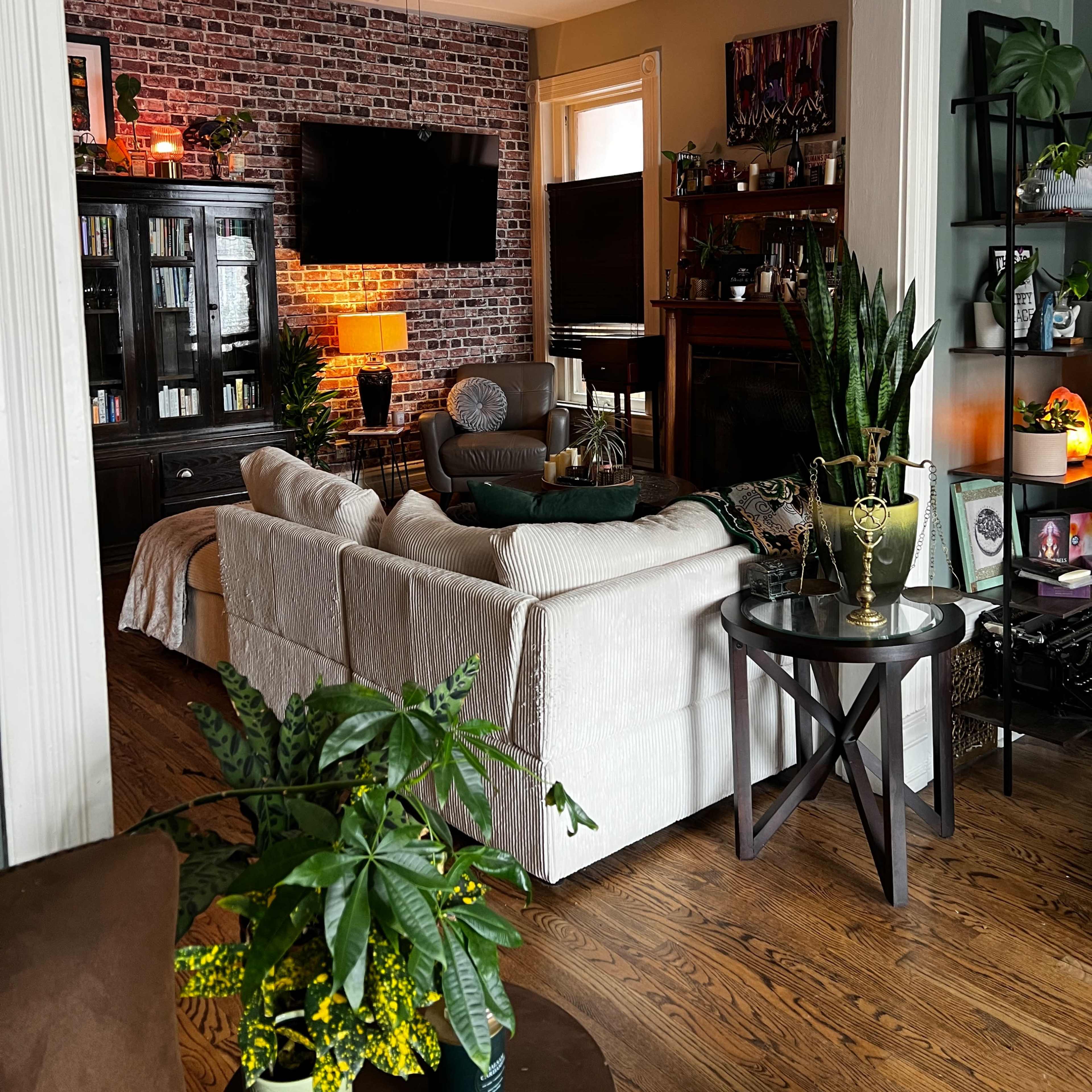 A cozy living room features a brick accent wall, a sectional sofa, a large television, and plants decorating the space.