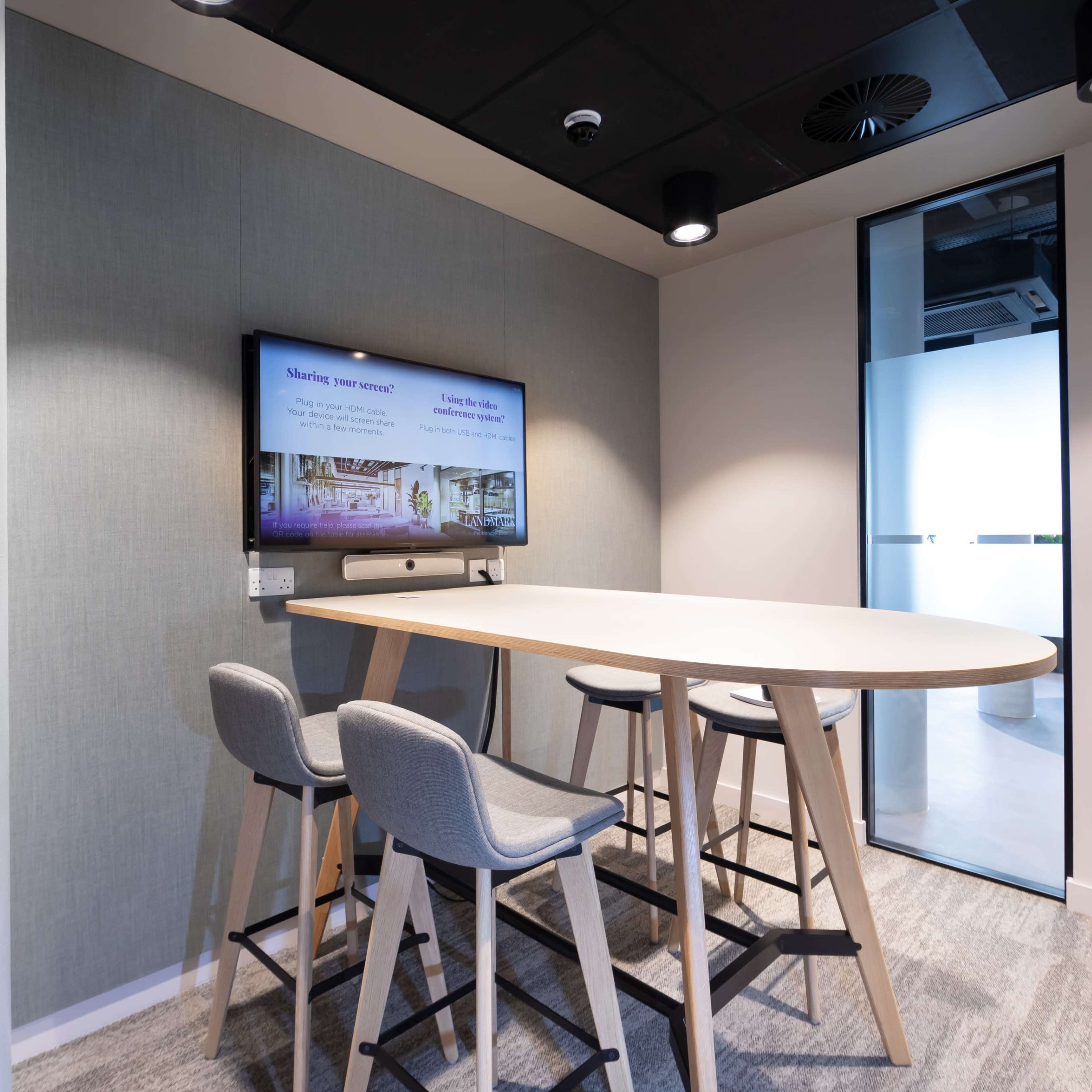 A small meeting room features a table with four stools and a wall-mounted screen displaying information.