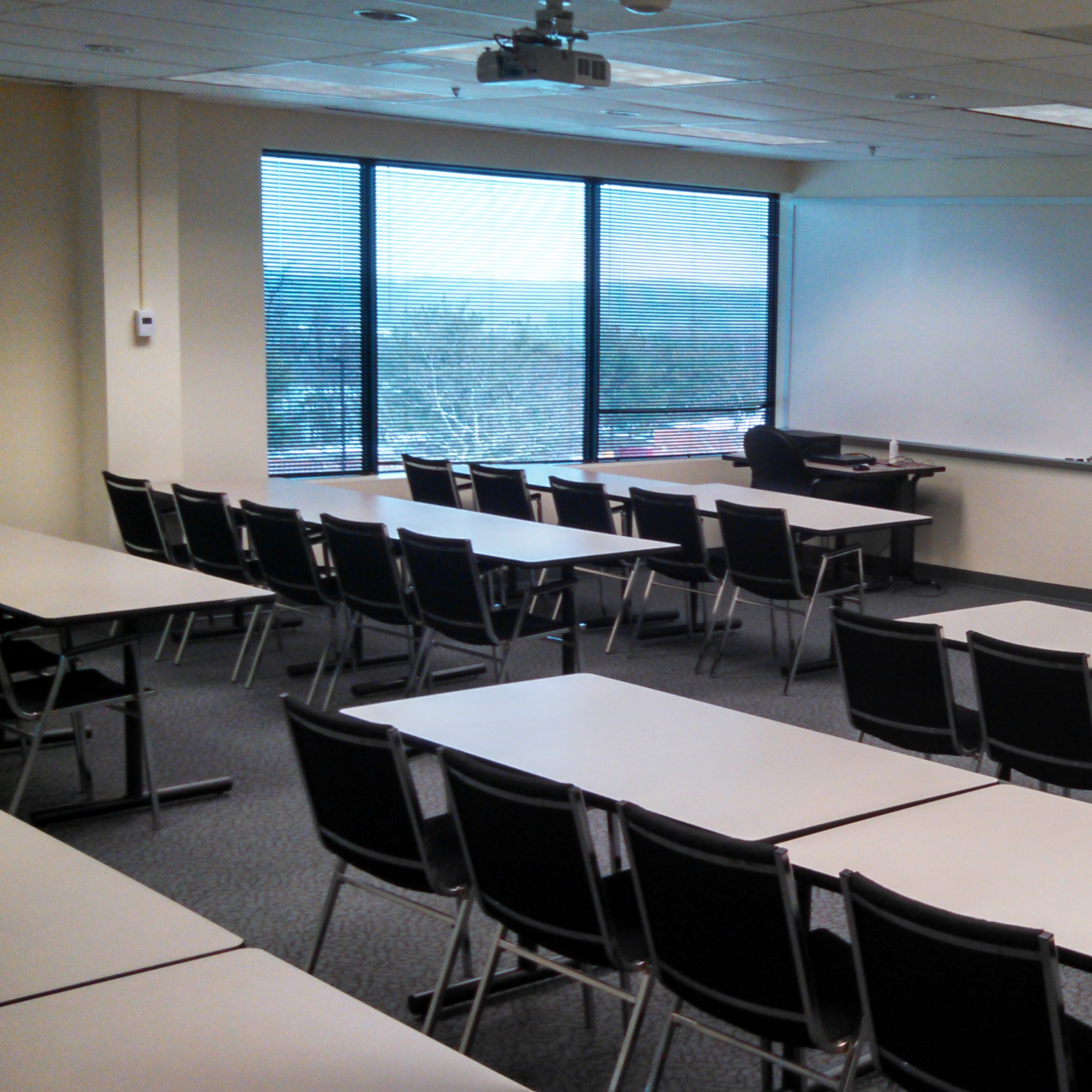 A classroom is arranged with several rows of tables and chairs facing a front whiteboard, with large windows providing a view of the outdoors.