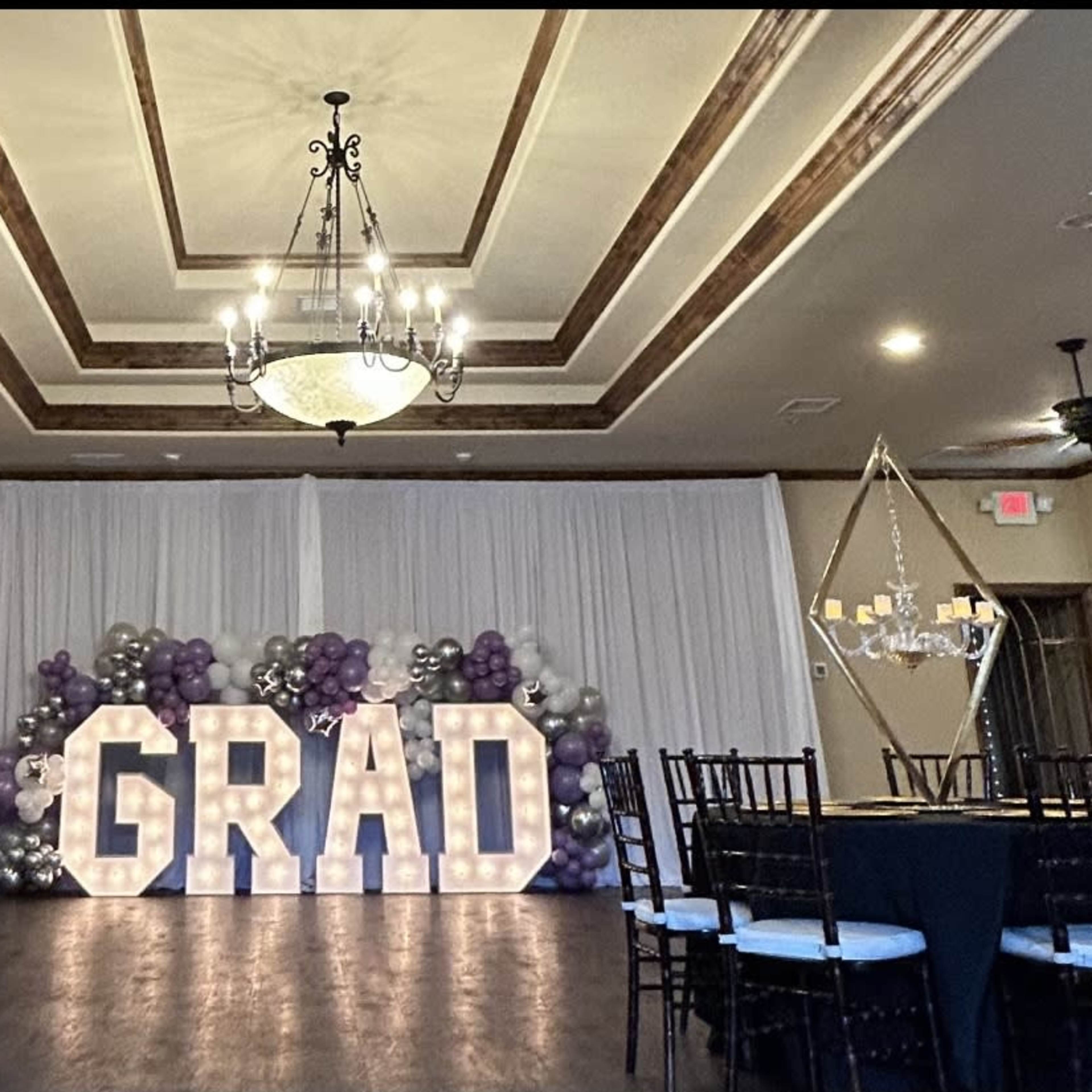 A banquet hall decorated for a graduation celebration, featuring large illuminated letters spelling "GRAD" against a backdrop of balloons in purple and silver.