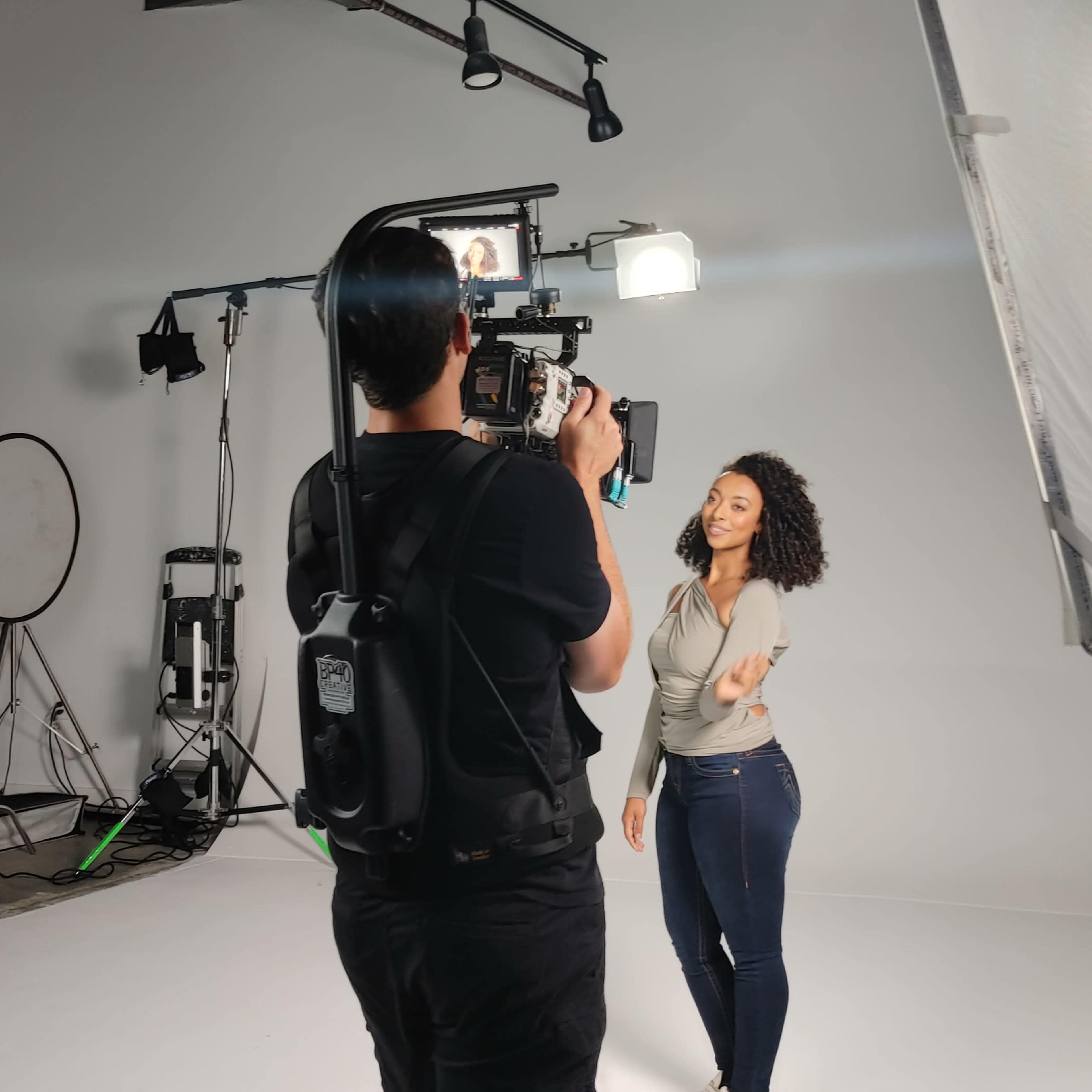 A photographer captures a subject posing in a studio with various lighting equipment and crew members in the background.