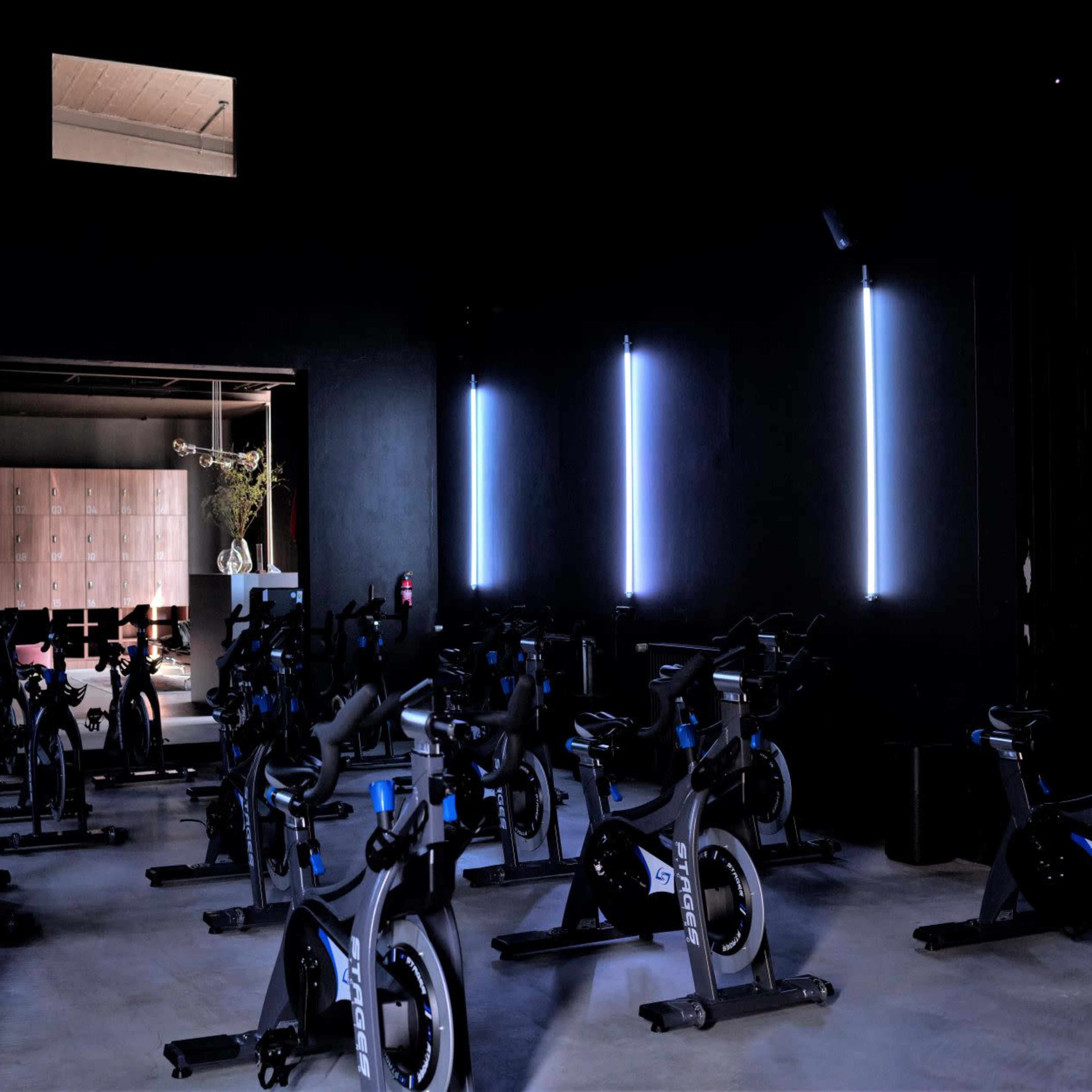 A dark indoor cycling studio features rows of stationary bikes illuminated by vertical strips of blue light.