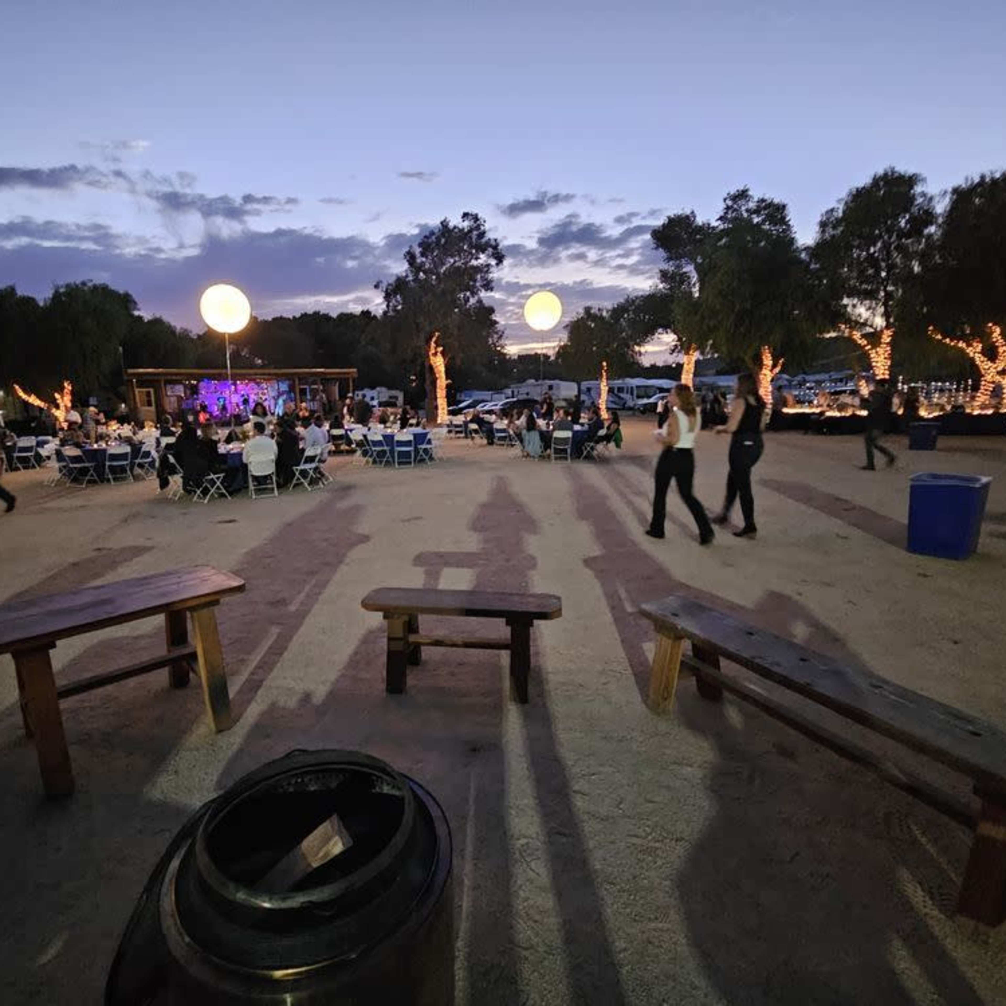A gathering is set up outdoors with tables and chairs under large illuminated balloons as dusk falls.