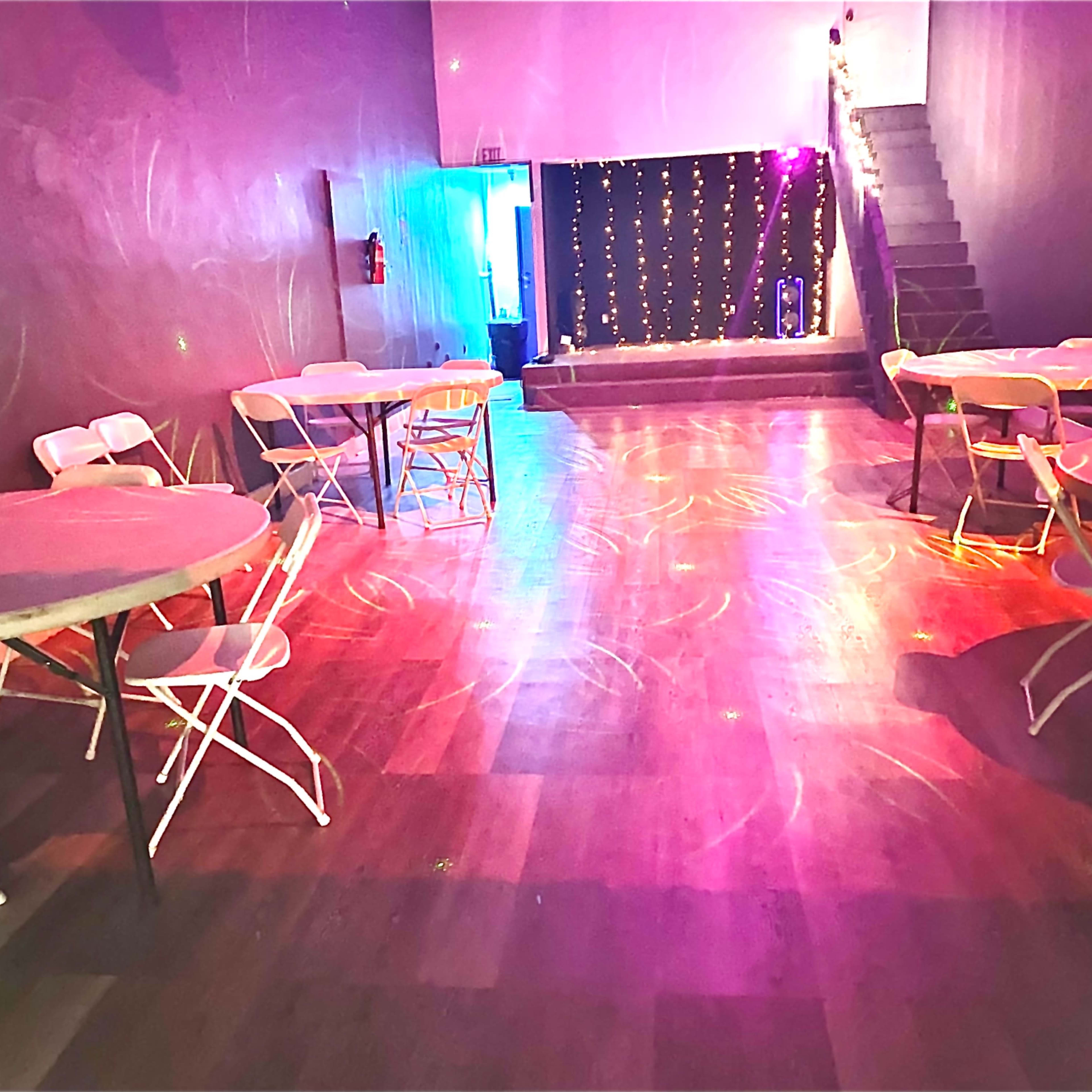 A dimly lit event space features several round tables, scattered chairs, and a small stage illuminated by colorful lights at the back.