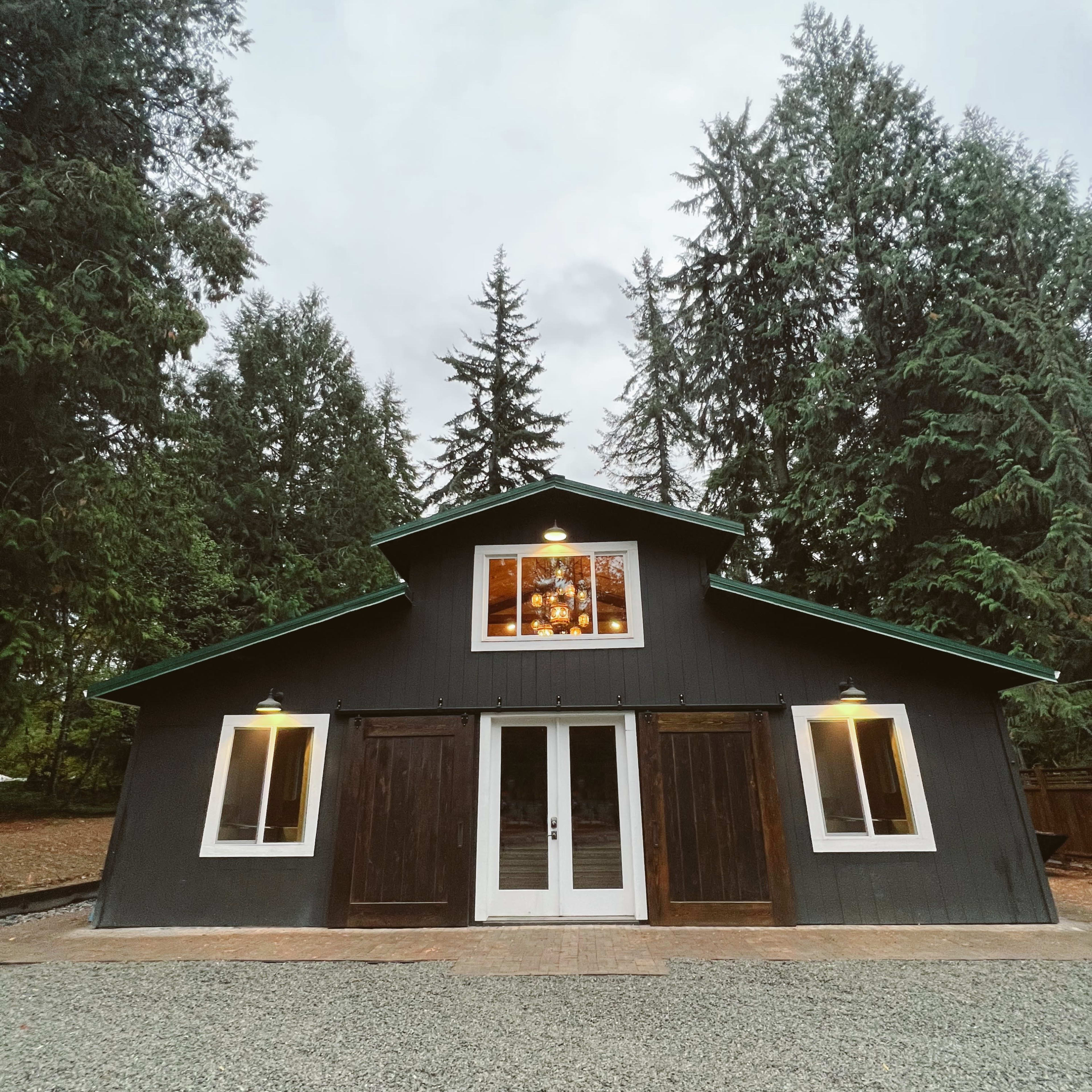 A dark-colored barn with large front doors and a windowed loft is surrounded by tall trees.