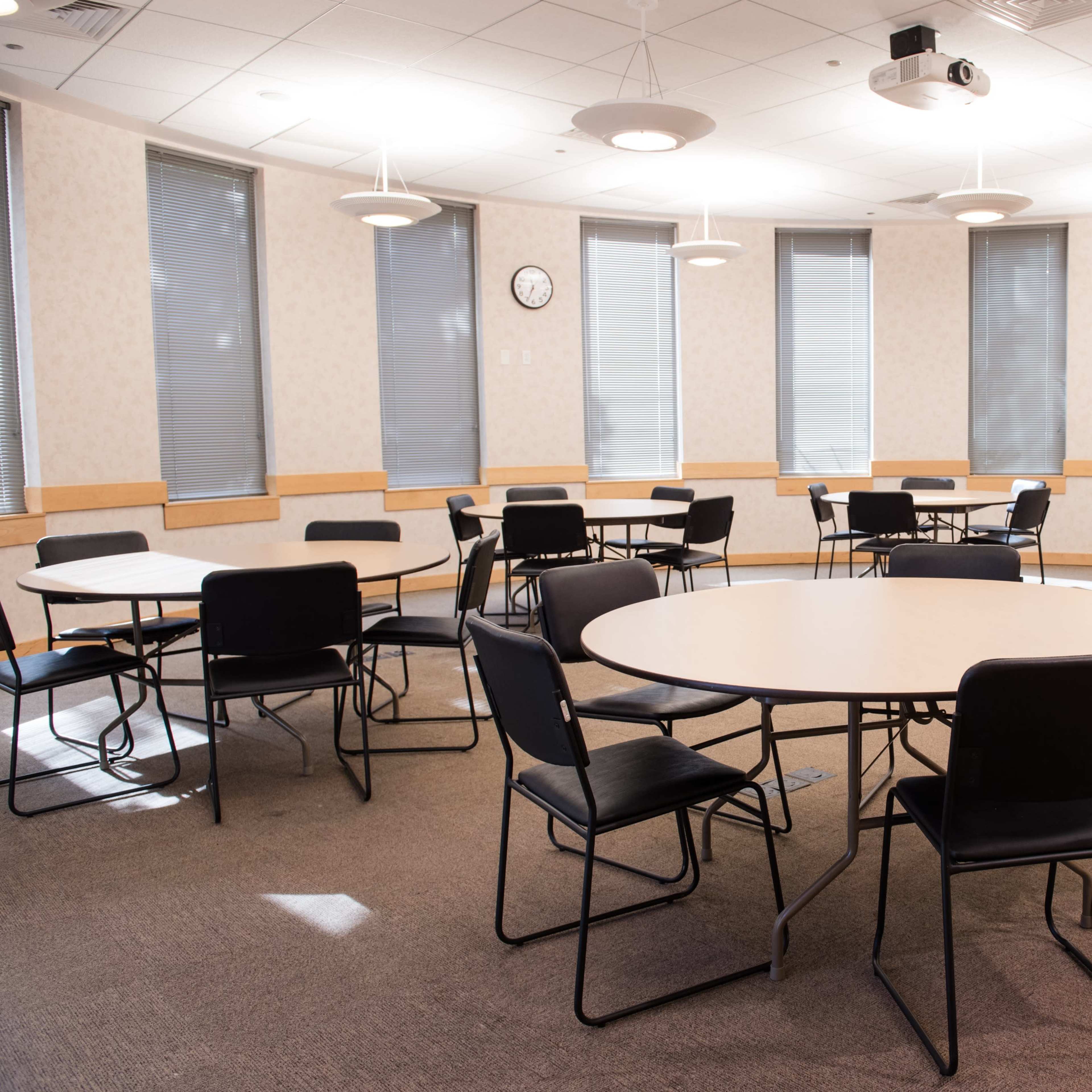 A mostly empty classroom with several round tables and black chairs arranged in a circular pattern under overhead lights.
