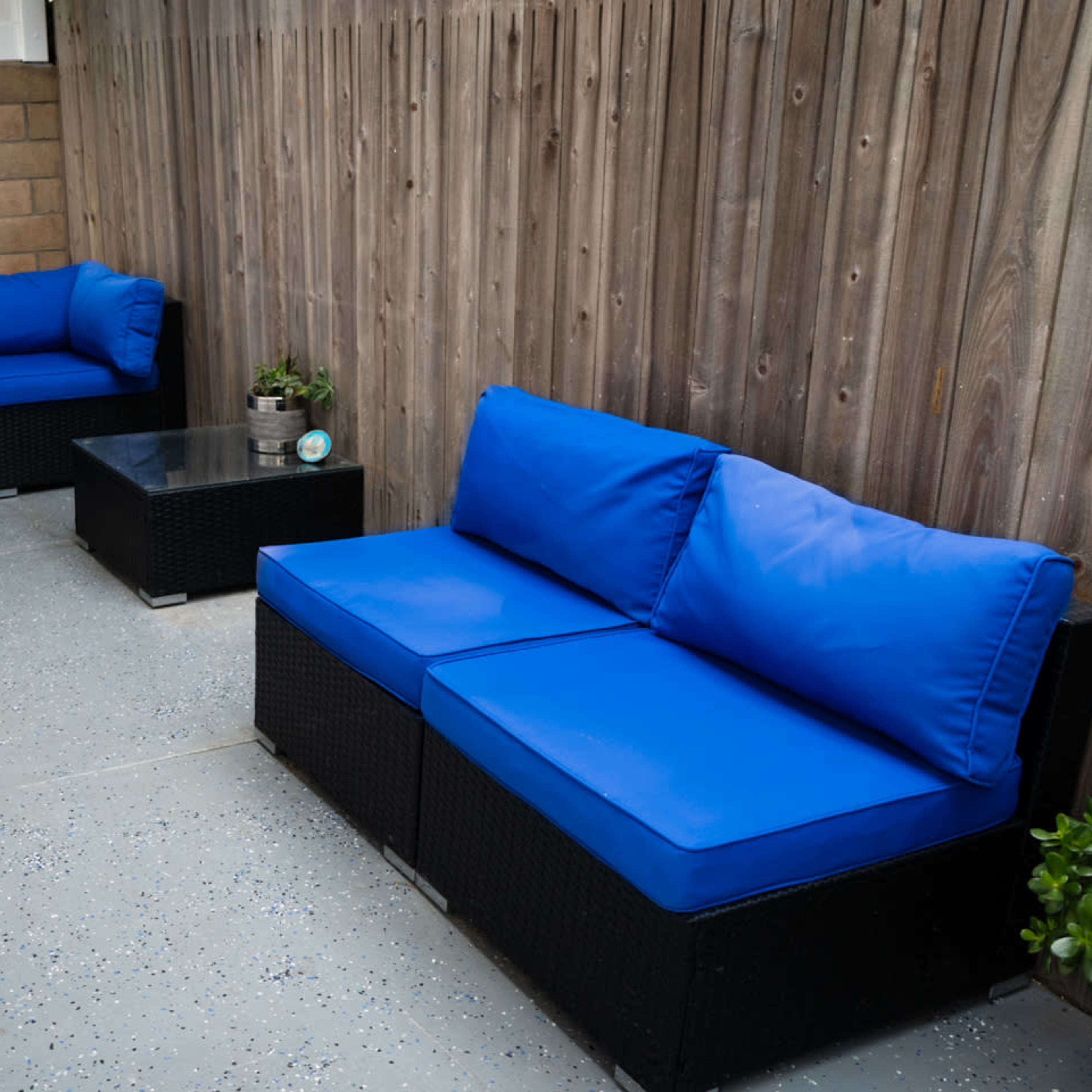 Two blue cushioned couches and a glass coffee table are arranged in a narrow outdoor space surrounded by a wooden fence.