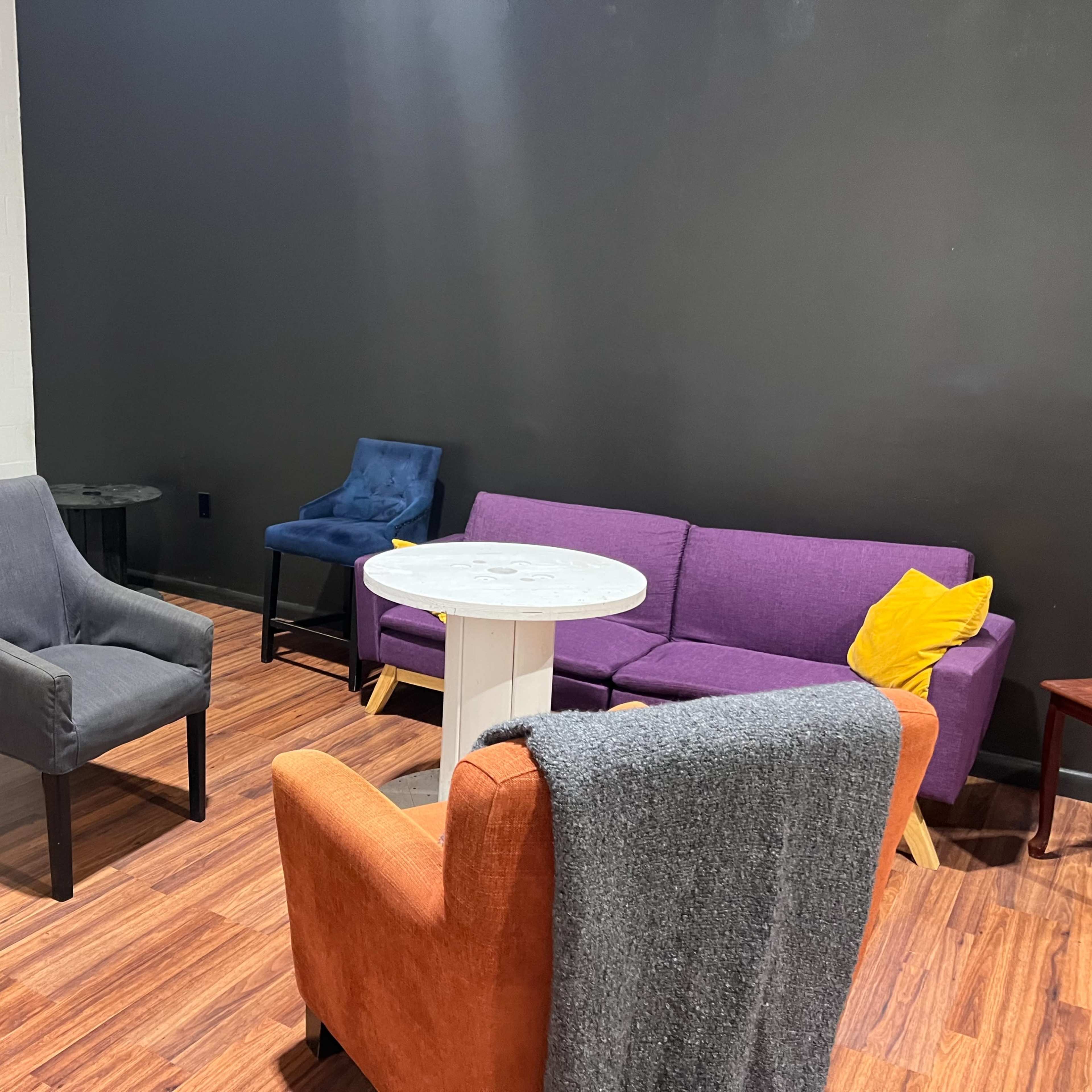 A seating area with a purple couch, an orange armchair, a gray armchair, a small white table, and a black side table against a dark wall.
