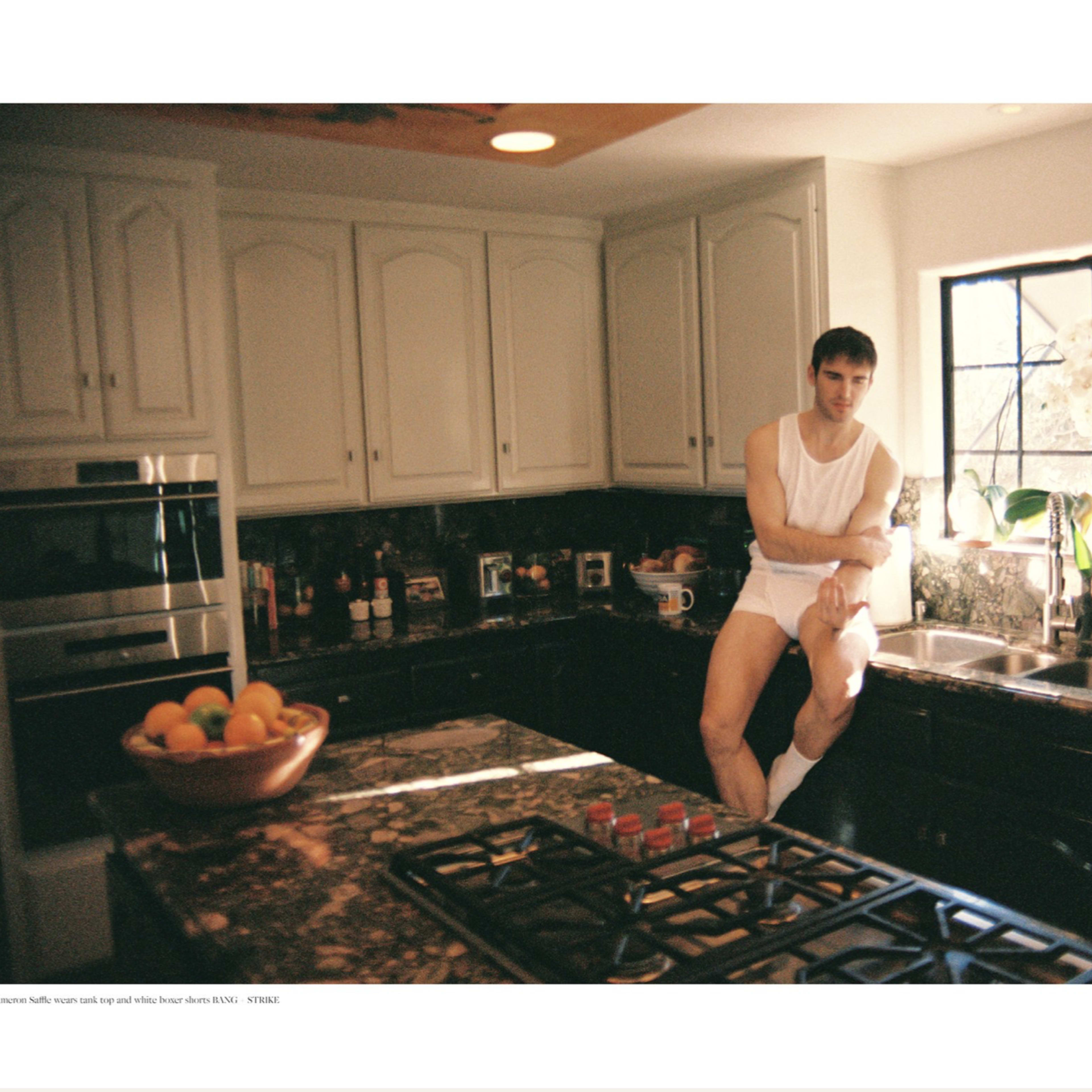 A person sits on the kitchen counter next to a sink in a home with white cabinetry and dark countertops.