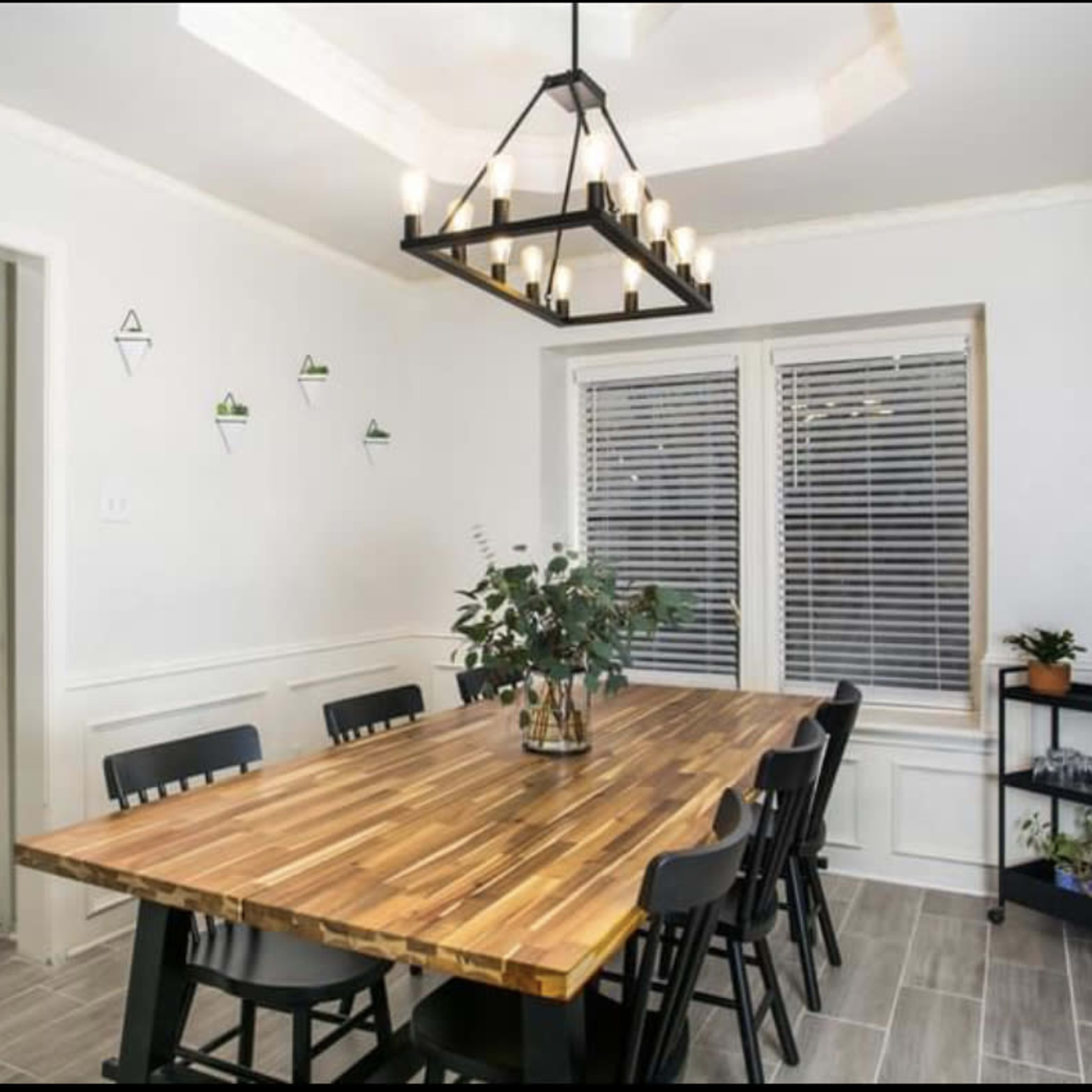 A dining area features a long wooden table surrounded by black chairs, with a modern chandelier overhead and plants decorating the space.