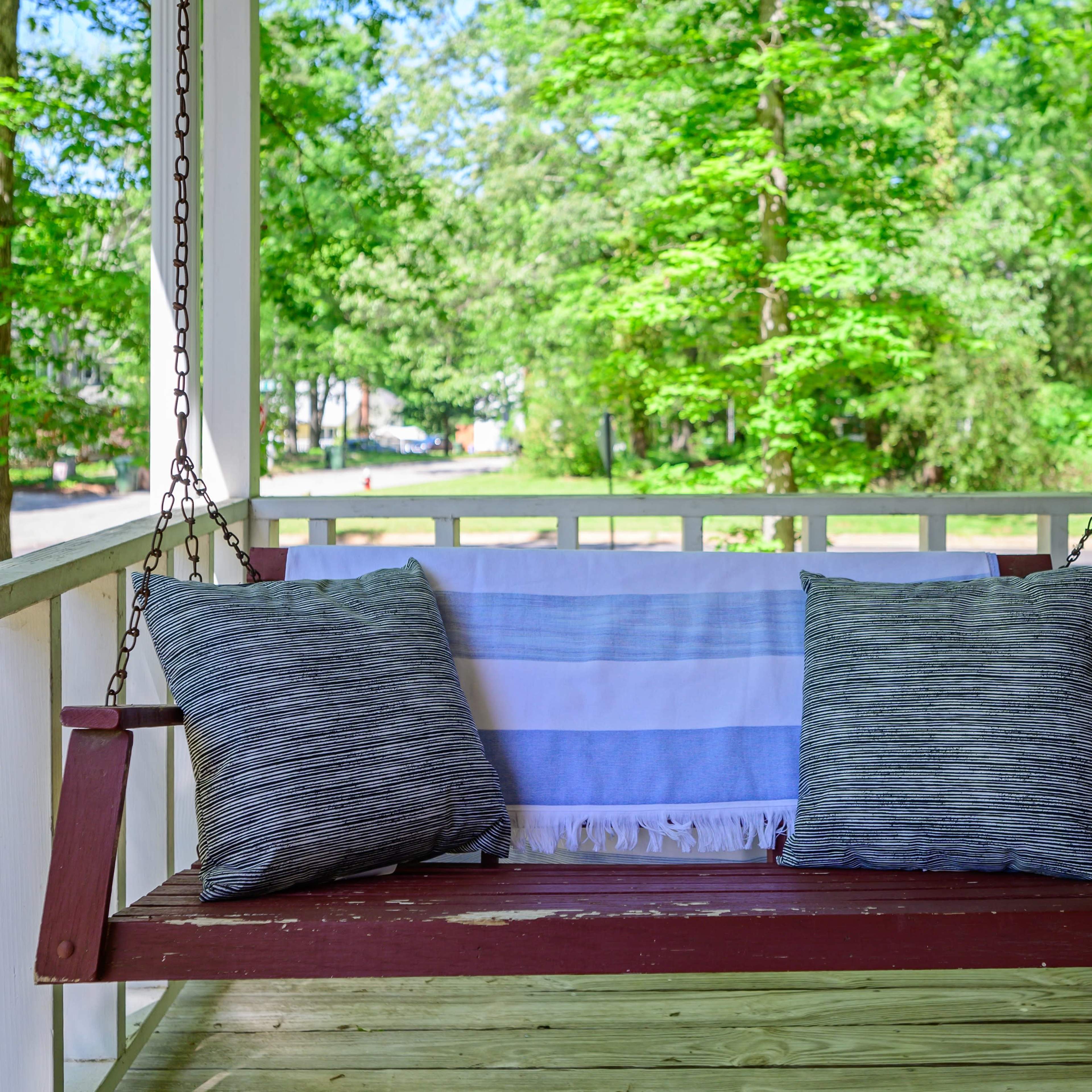 A wooden porch swing with two pillows is positioned on a porch overlooking a green, tree-filled yard.