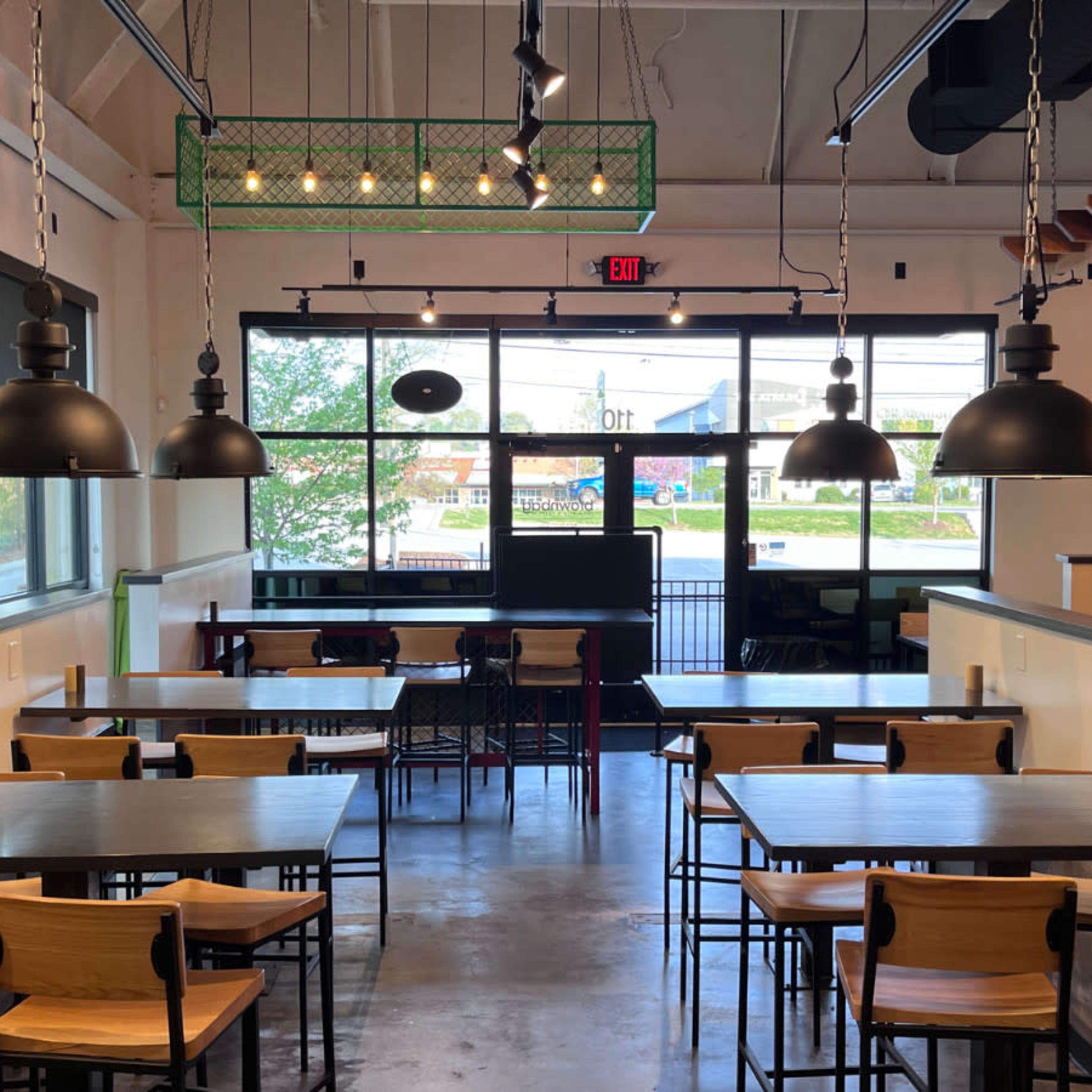 A modern restaurant interior with a series of wooden tables and chairs, large pendant lights, and a glass entrance door.