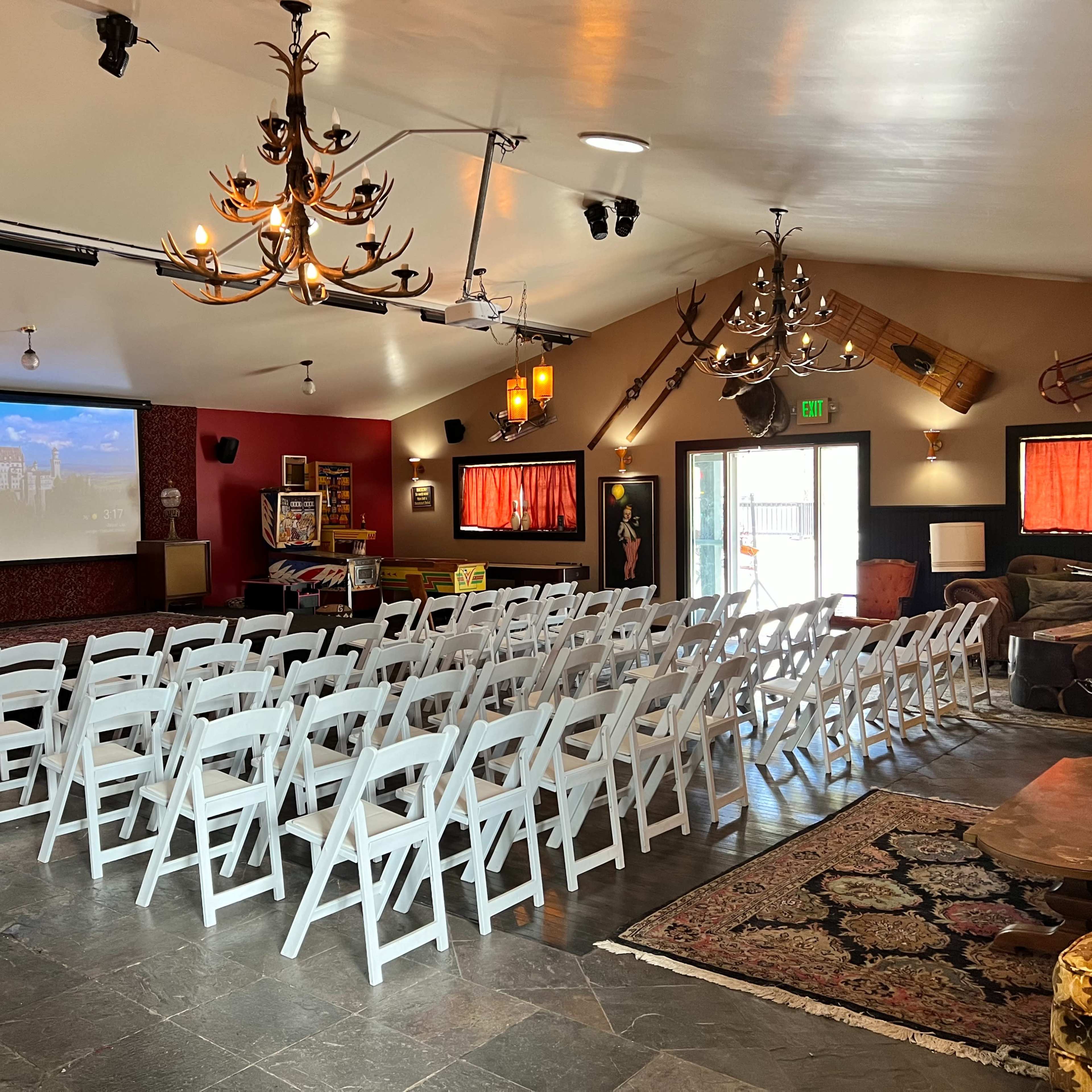 A spacious indoor venue set up with rows of white folding chairs facing a large projection screen, with chandeliers and decorative elements on the walls.