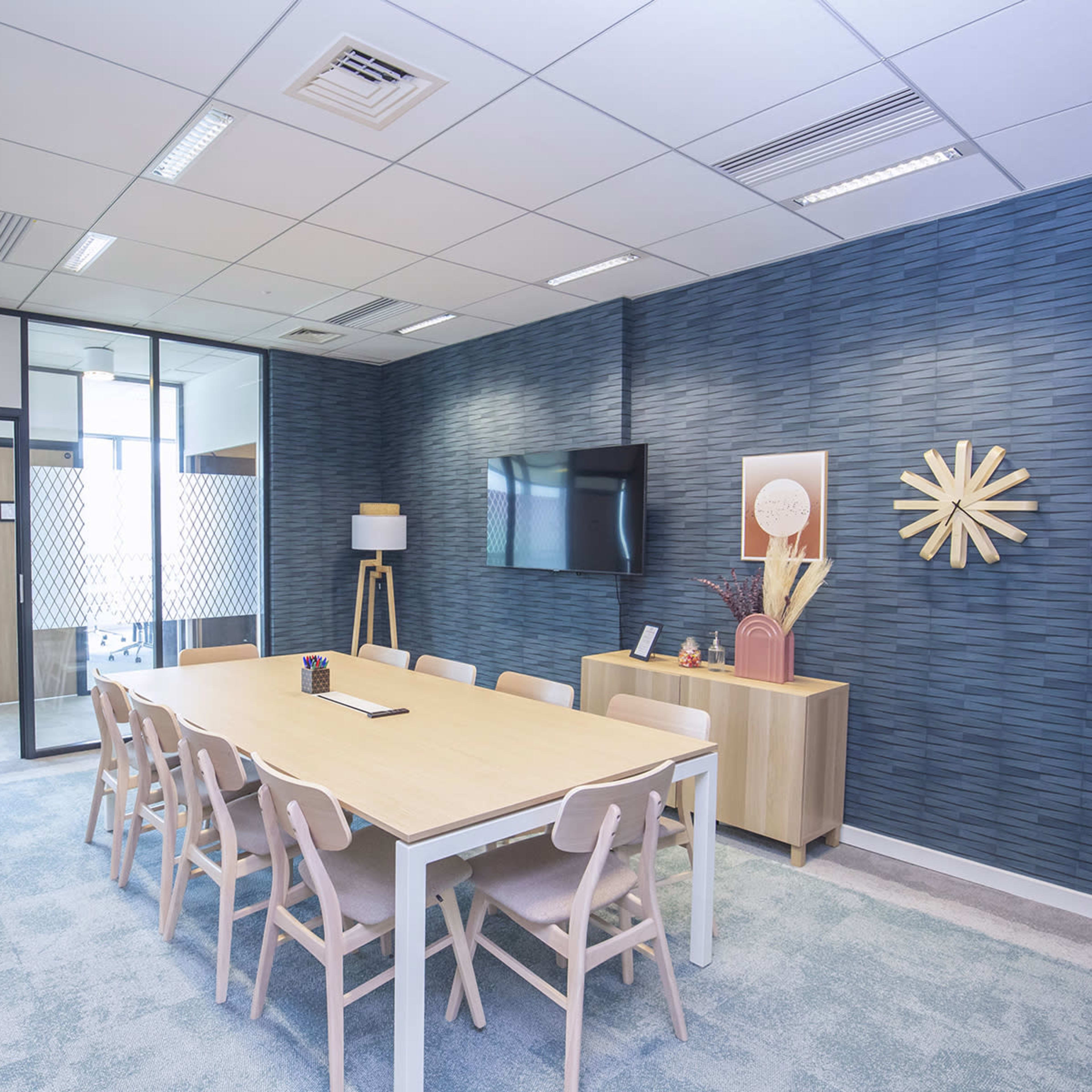 A conference room features a long table surrounded by chairs, with a blue textured wall, a television, and decorative elements.
