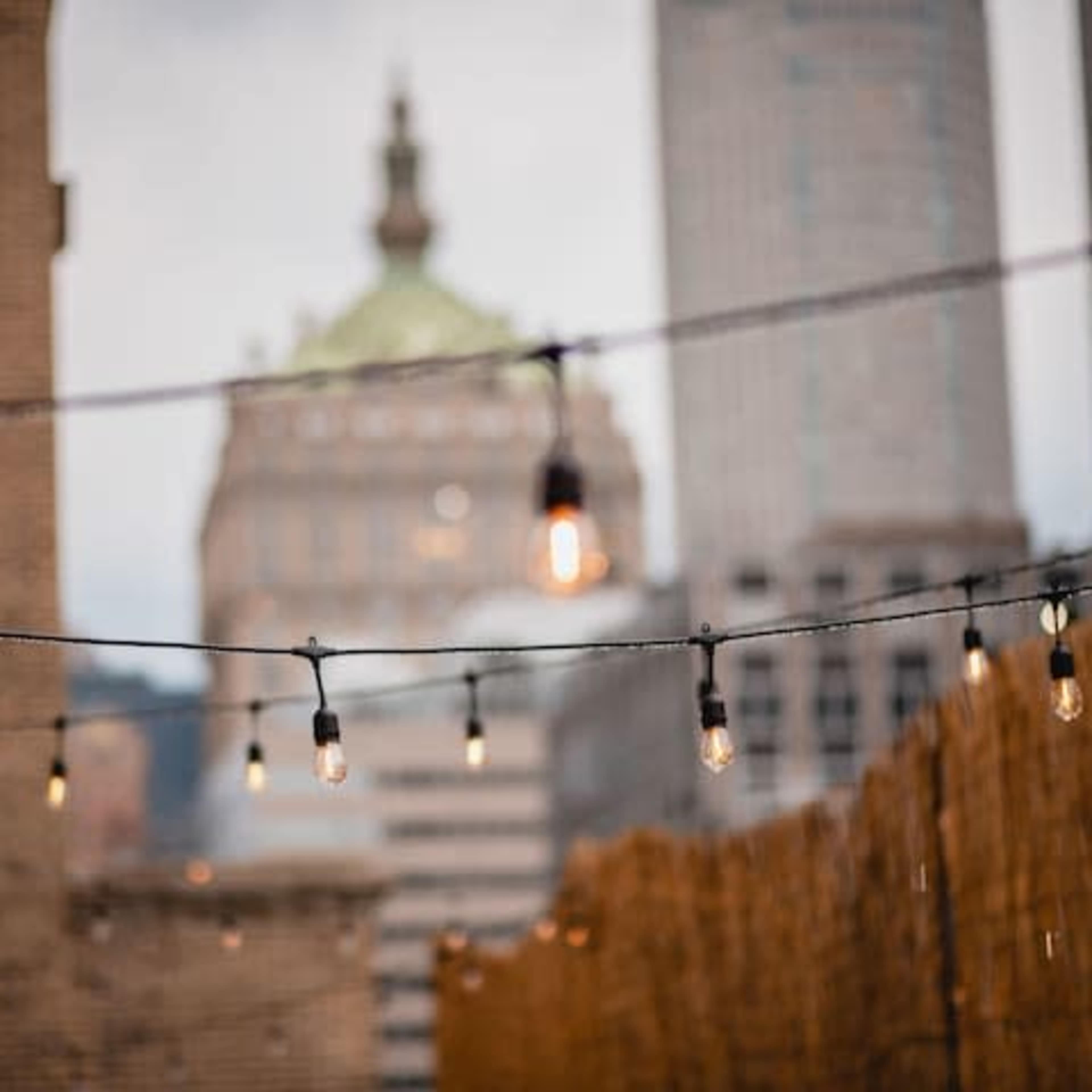 String lights are strung across a rooftop, with a city skyline and a dome building visible in the background.