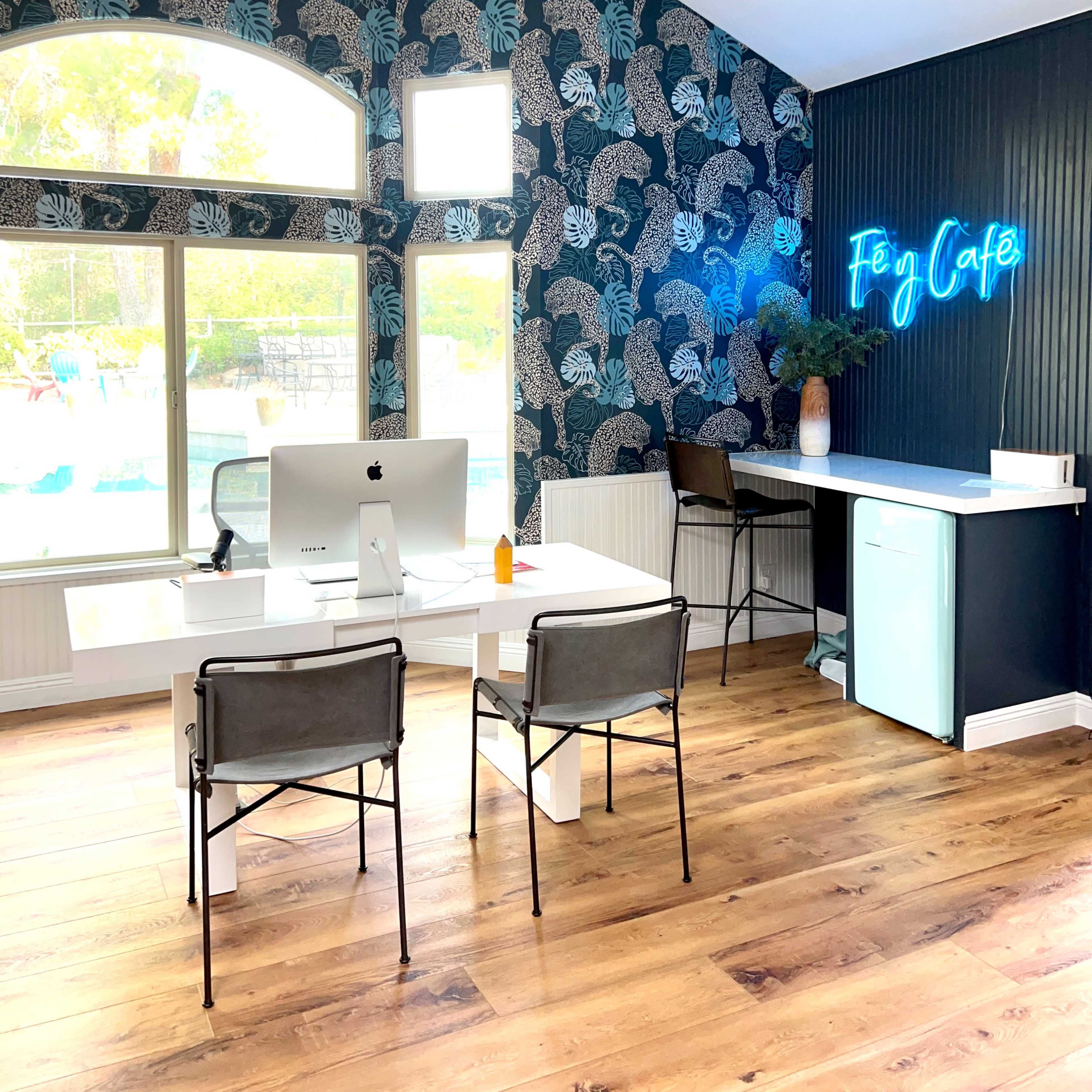 A modern home office features a large desk with an Apple computer, two chairs, a mini fridge, and a vibrant floral wallpaper.