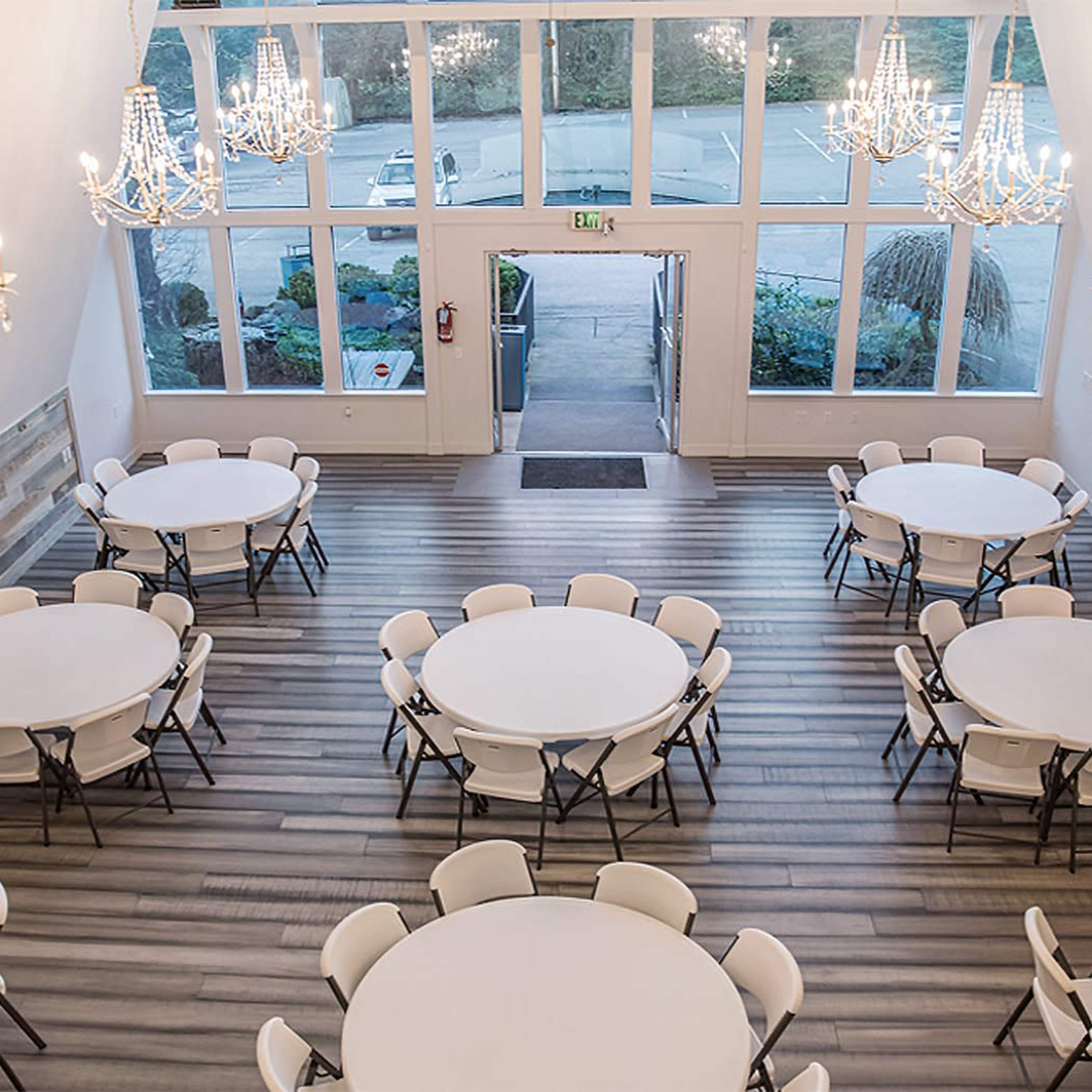 A spacious banquet room with round tables set up and large chandeliers hanging from the ceiling, illuminated by natural light from large windows.