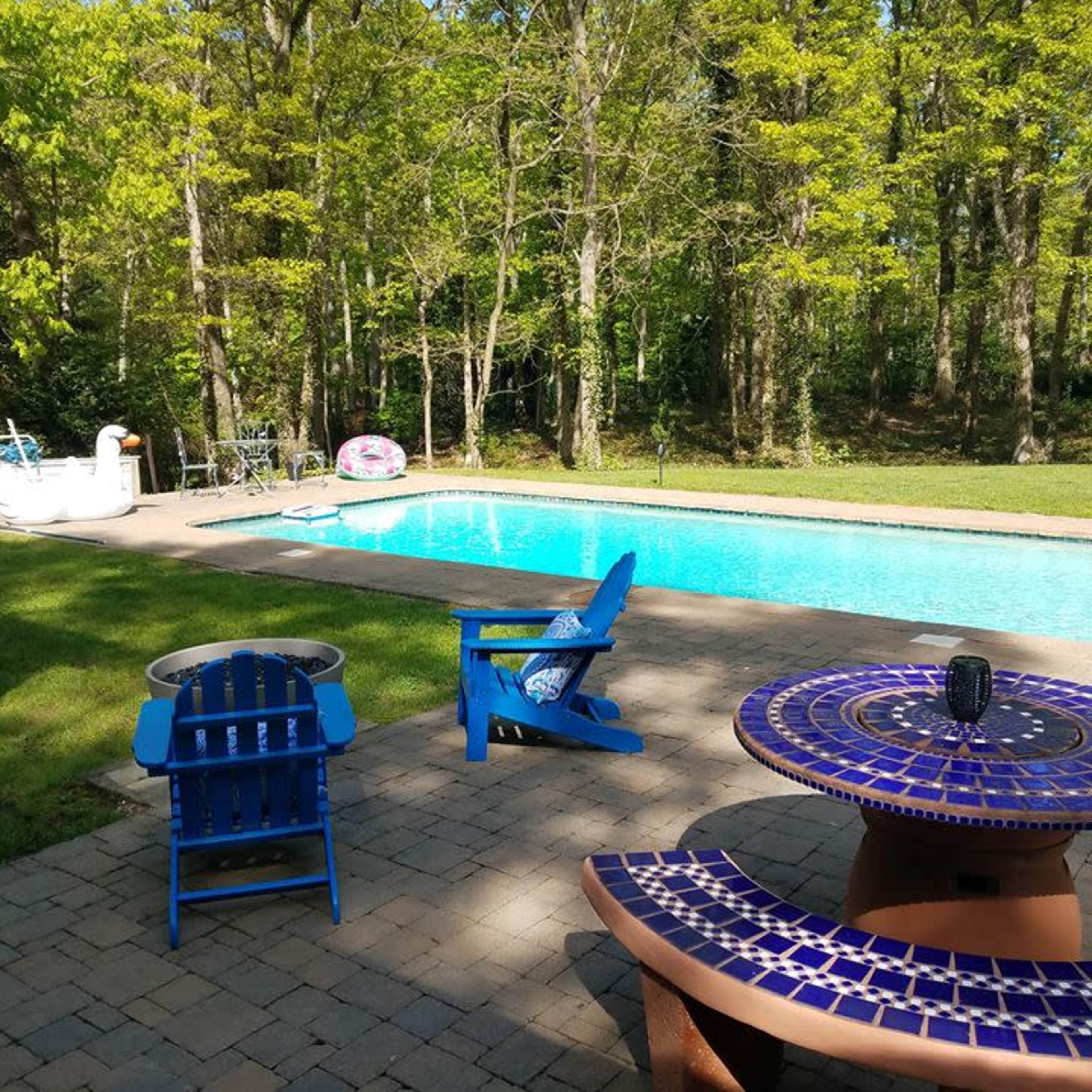 A backyard scene features a blue pool surrounded by trees, with colorful patio furniture and a stone patio.