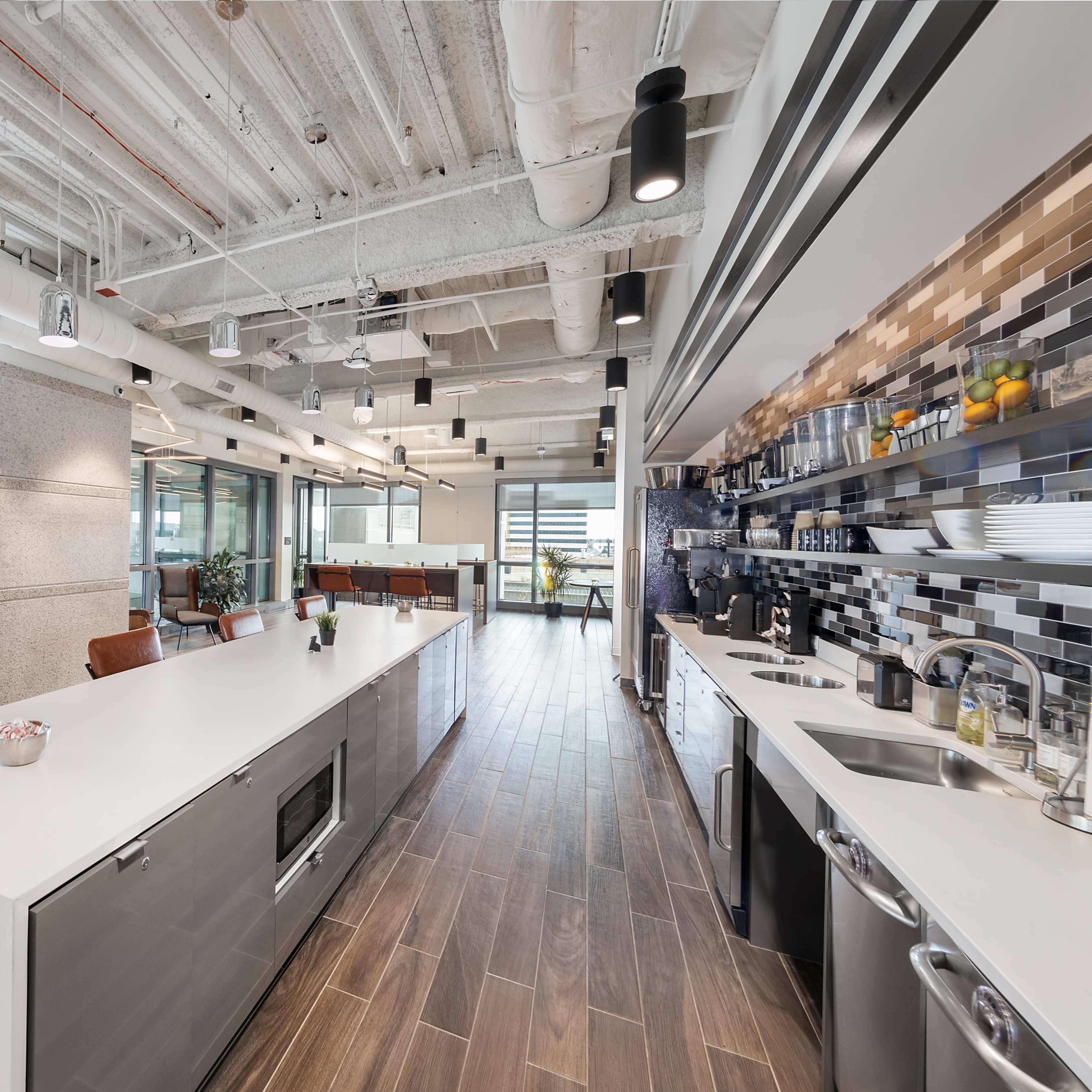 A modern kitchen and dining area in an office space, featuring an elongated countertop, appliances, and seating arrangements.