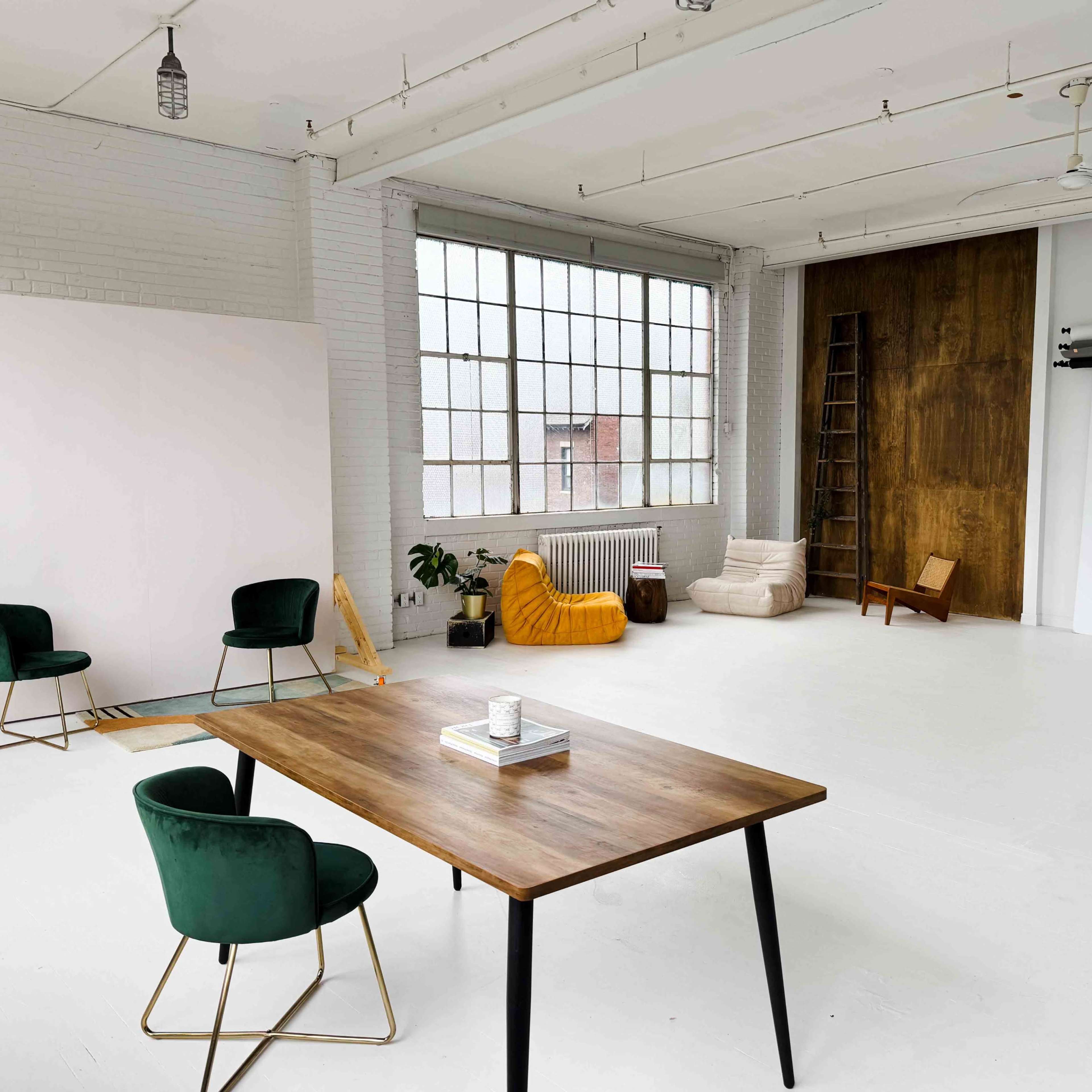 A spacious, well-lit studio features a wooden table, green chairs, a yellow beanbag, and a white backdrop near large windows.