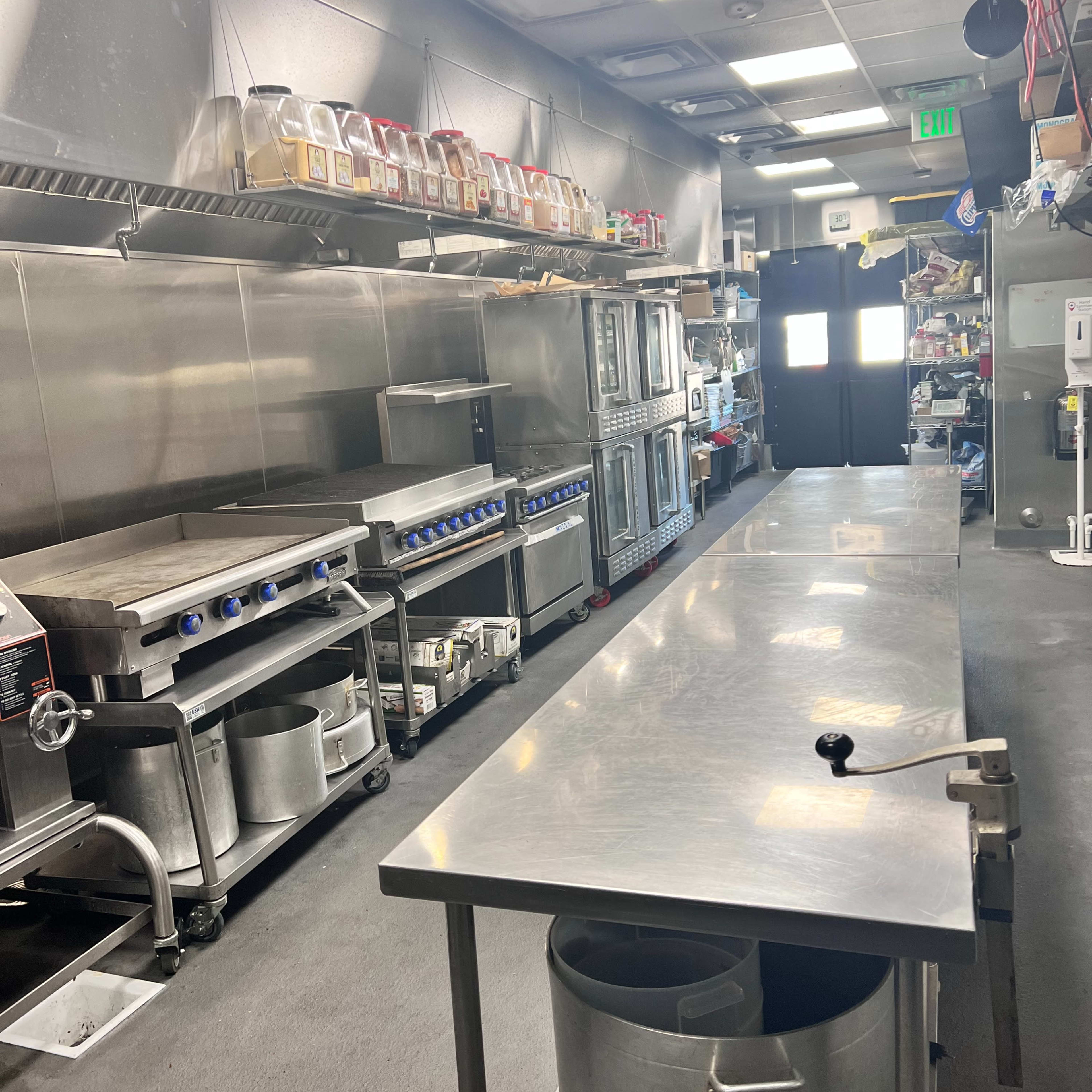 A commercial kitchen equipped with stainless steel appliances, a long prep table, and storage shelves filled with various containers.