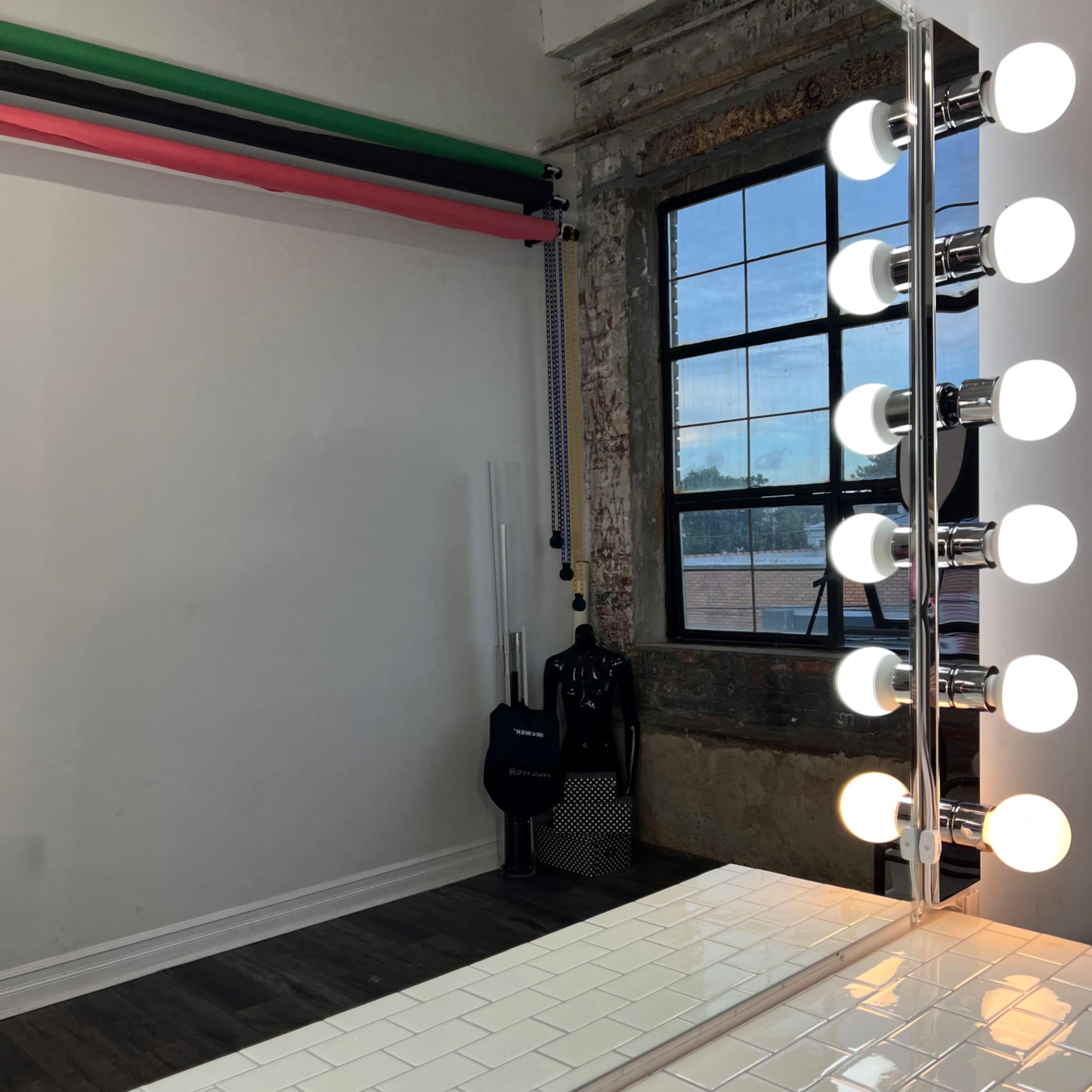 A well-lit vanity area with a large mirror and bright round bulbs, set against a wall with colorful backdrop supports and a large window.