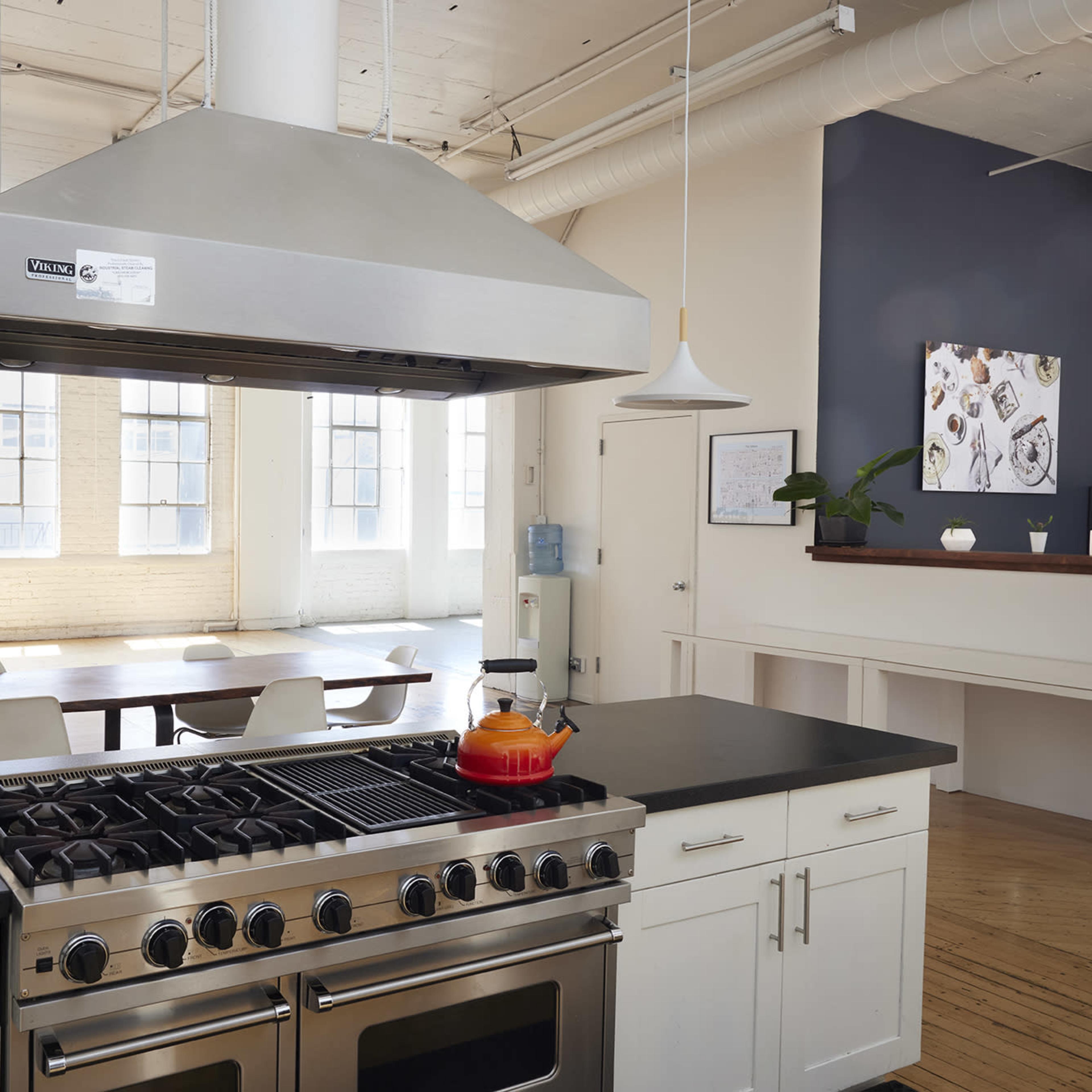 A modern kitchen with a stainless steel hood, a gas stove, and a dining area featuring a long table and chairs, illuminated by natural light from large windows.