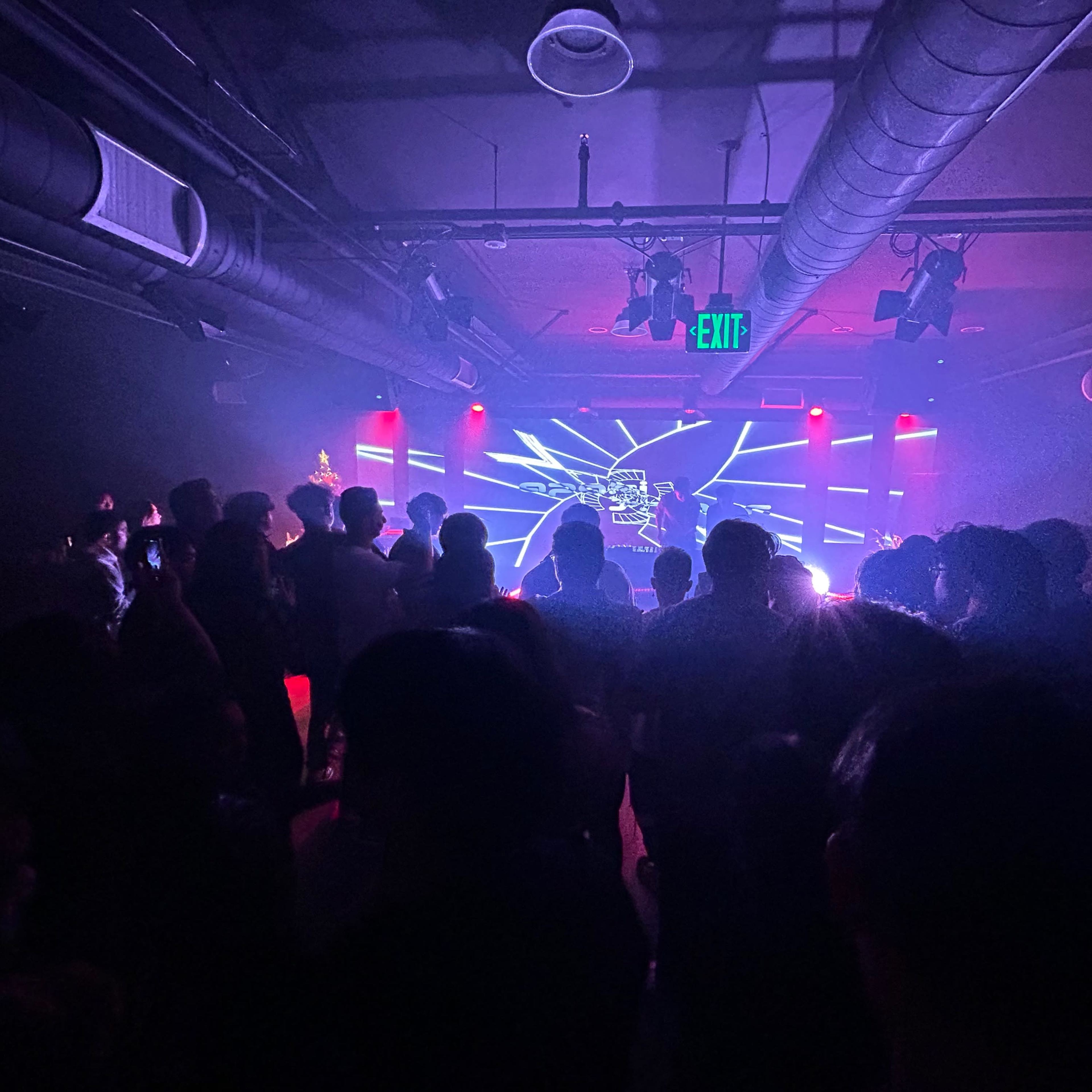 A crowd gathers in a dimly lit venue with colorful lights and visual displays illuminating the stage.