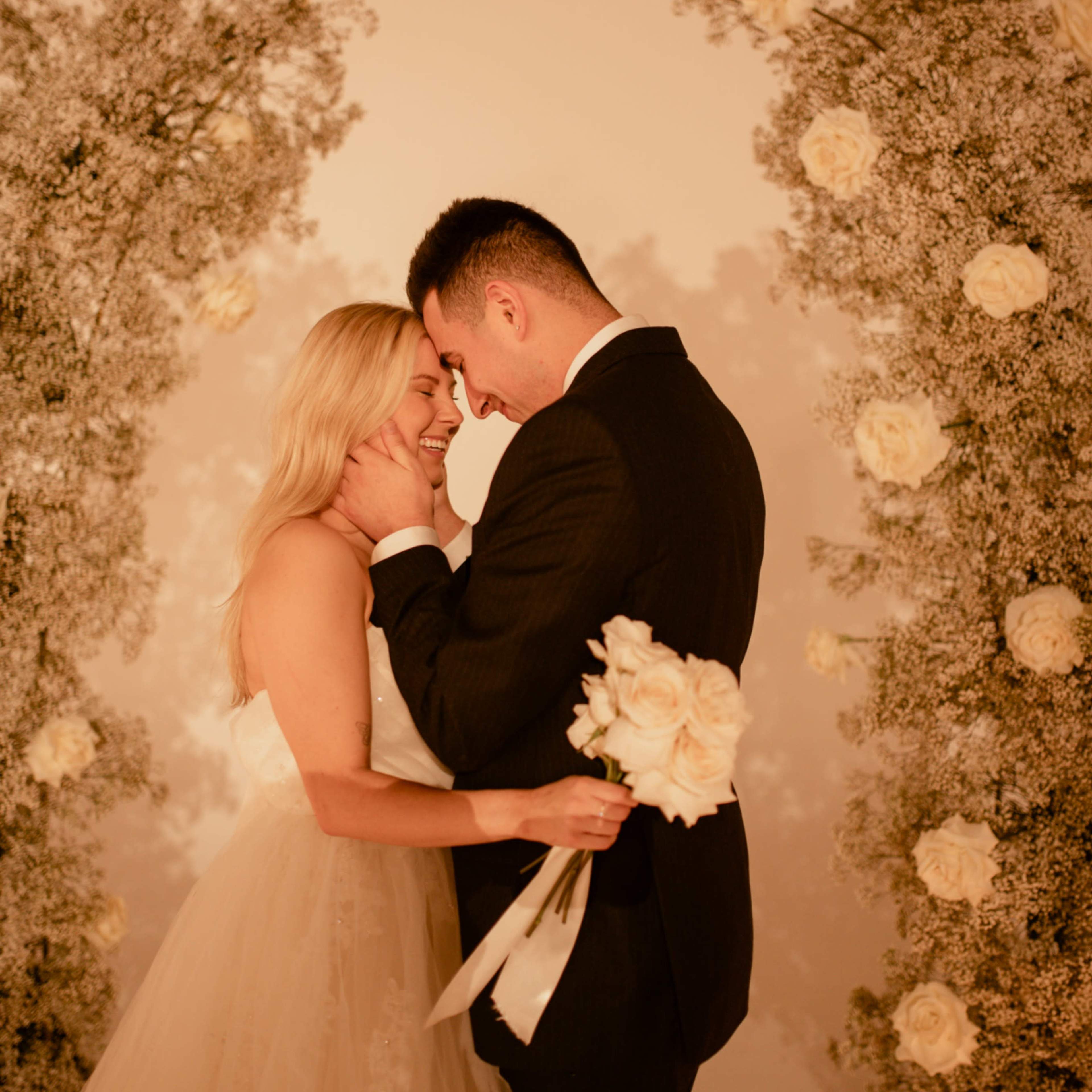 A couple stands close together, smiling and holding a bouquet of roses, with floral arrangements in the background.