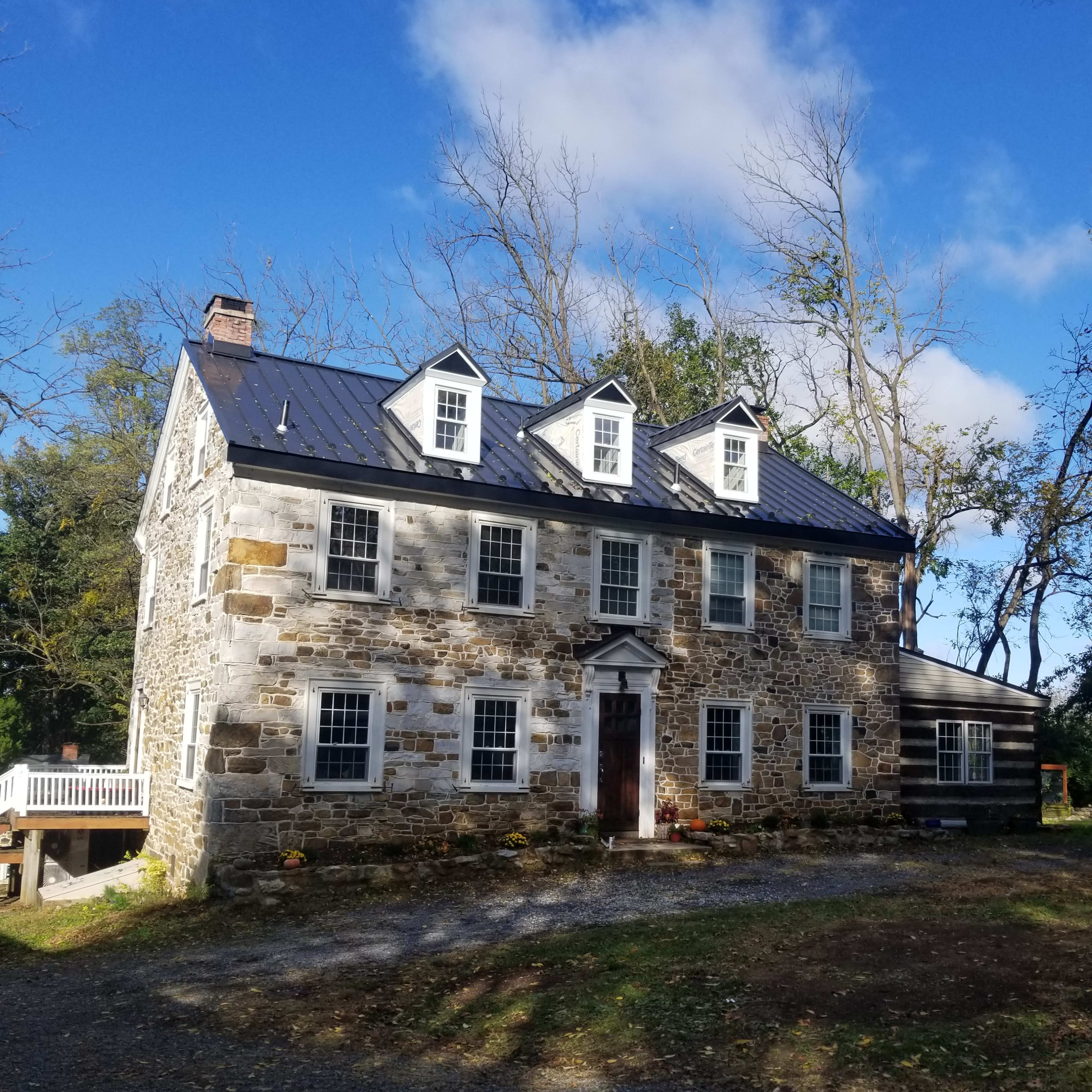 A stone house with a slate roof and multiple windows stands amidst a gravel driveway and trees under a partly cloudy sky.