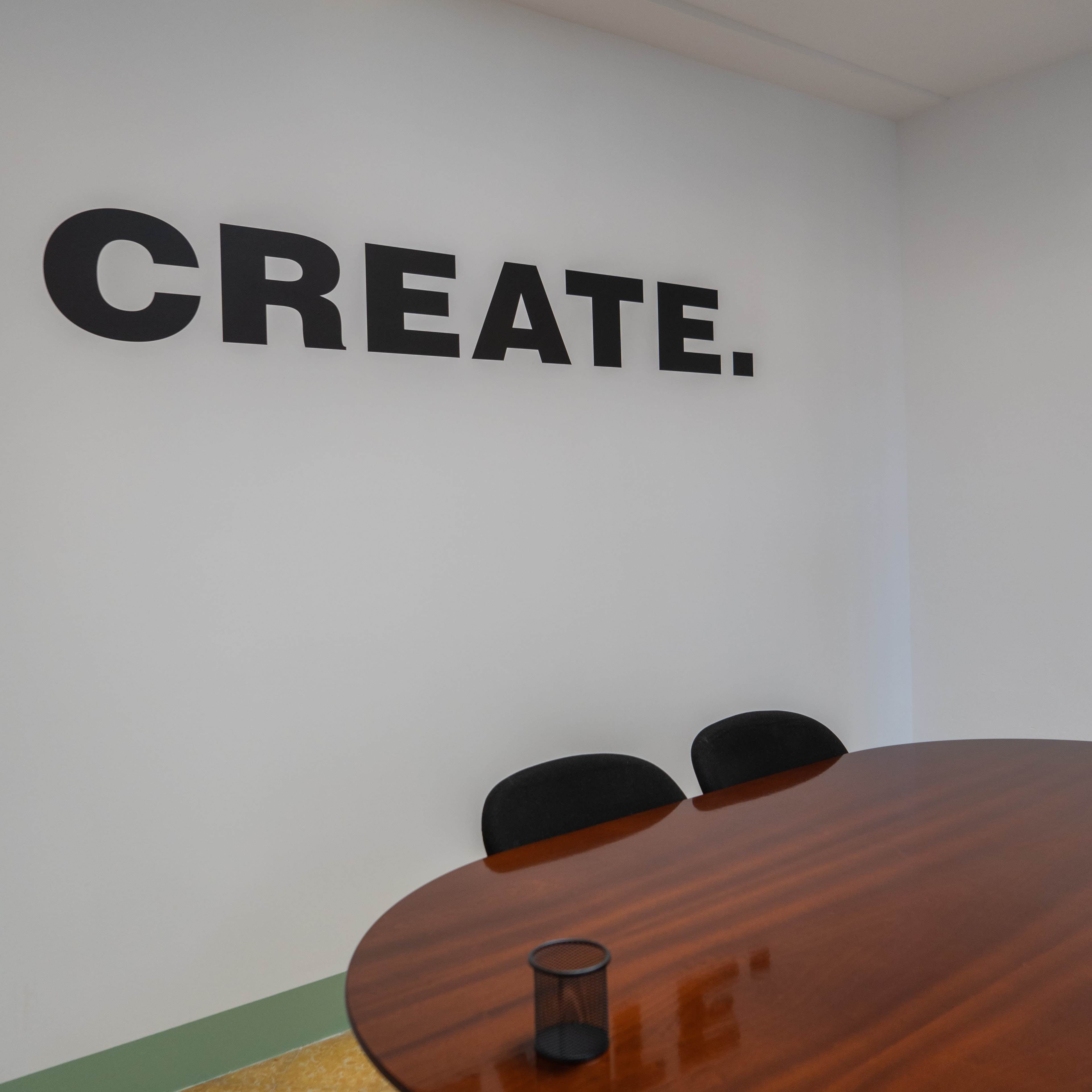 A round wooden table with black chairs is situated in a room featuring a large wall sign that reads "CREATE."